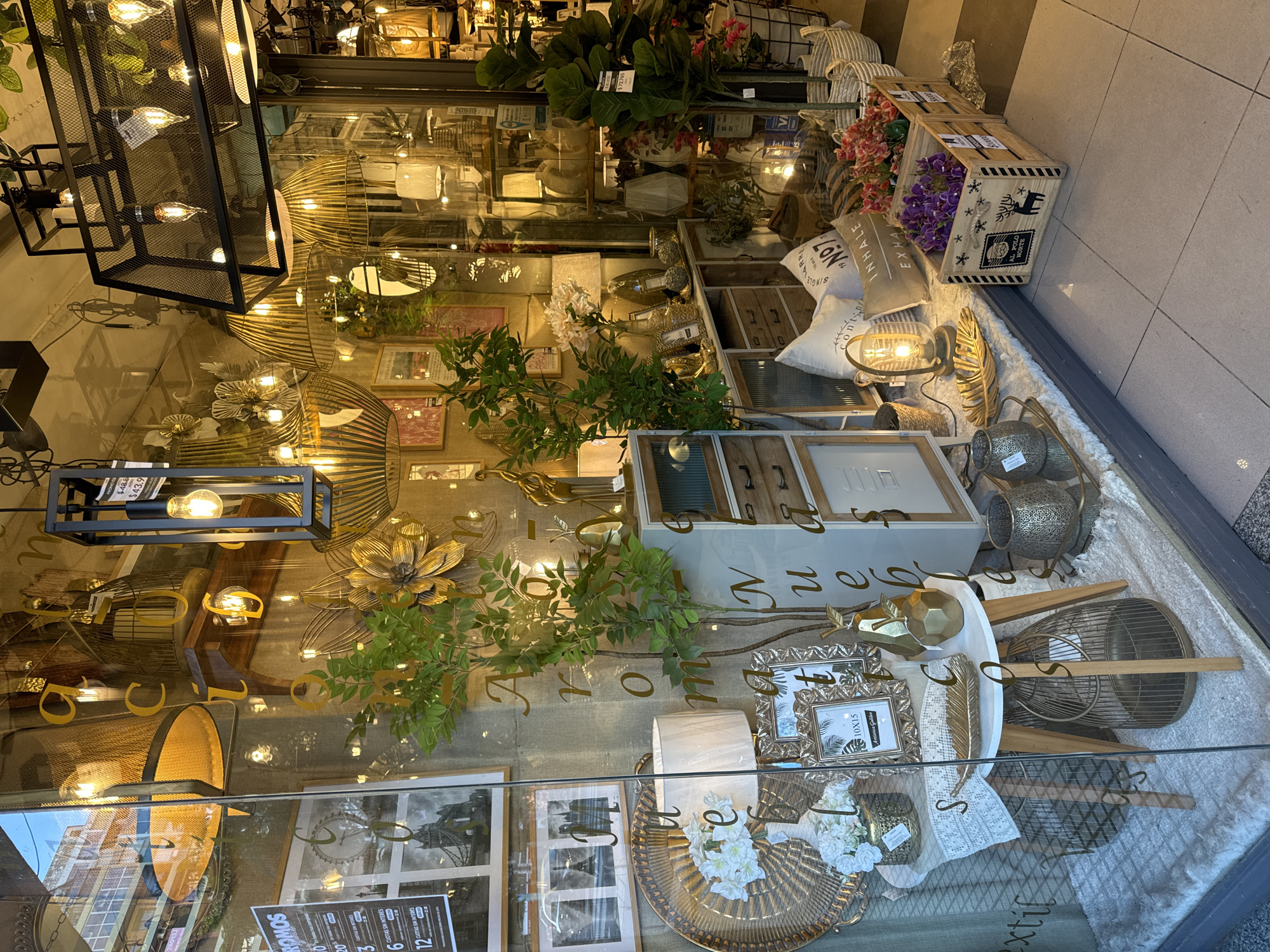 The image captures a detailed and visually rich retail display window, rotated 90 degrees counter-clockwise. The setting is likely a home decor or gift shop, showcasing a diverse collection of decorative items with a predominant palette of gold, black, and natural earthy tones.
The display is packed with various objects. Numerous decorative light fixtures are central, featuring exposed Edison-style bulbs within black metal frames and golden, cage-like structures. Artificial plants with green foliage and arrangements of purple, pink, and white flowers add natural accents throughout. A rustic wooden crate, adorned with a reindeer motif and the text "AL POLO NORTE," holds some of these floral arrangements. Other decor includes decorative pillows with text such as "SINGLE," "INHALE," and "EXHALE," framed pictures (some depicting the Eiffel Tower), a golden woven tray, and various metallic sculptures including a large golden flower and geometric shapes. A fluffy white textile or rug covers the base of the display, creating a soft foundation.
The store's interior is brightly lit with warm artificial light, highlighting the textures and metallic sheen of the items. The glass window reflects elements of the outside environment, including a building with windows, overhead lights, and the pavement, creating a layered and somewhat distorted view of both the interior and exterior.
Visible text includes what appears to be a brand name, "SOWON," along with price tags such as "$72.765," "$48.8," and "$43.9." Advertisements for "6" and "12 CUOTAS SIN INTERES" (installments without interest) are prominently displayed, a common payment option in Argentina. Other readable words include "PROM" (promotion), "Nuebl" (likely part of 'Muebles' for furniture), "e extil" (likely 'Textil' for textile), and "10X15" on a photo frame. No people are visible, and the scene depicts a static, carefully arranged retail presentation.