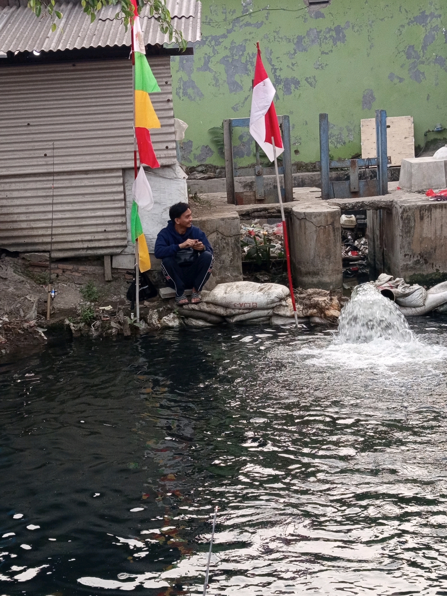A man is sitting on sandbags by a body of water in Karawang, Indonesia. He is wearing a dark blue tracksuit and is holding a phone. In front of him, there are a few fishing rods. Behind him, there is a corrugated metal building and flags. In the water, there is a fountain of water. The environment appears to be an urban area.