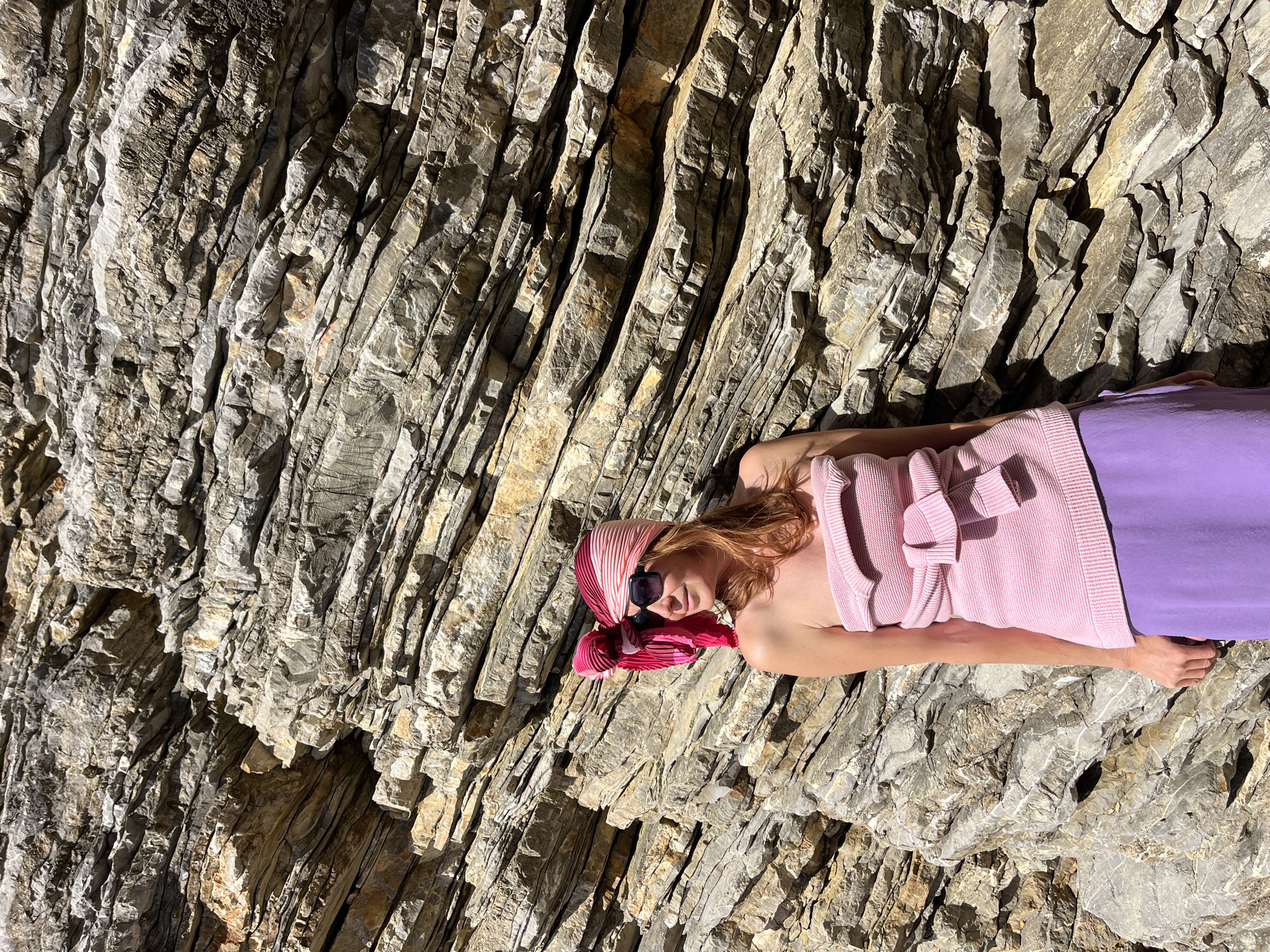 A woman lies horizontally against a geological rock formation. The woman is wearing sunglasses, a red and pink patterned scarf on her head, and a pink sweater tied around her chest. Her lower body is clad in a purple garment. The rock formation behind her is composed of grey and brown layers. The location is Budva, Montenegro.