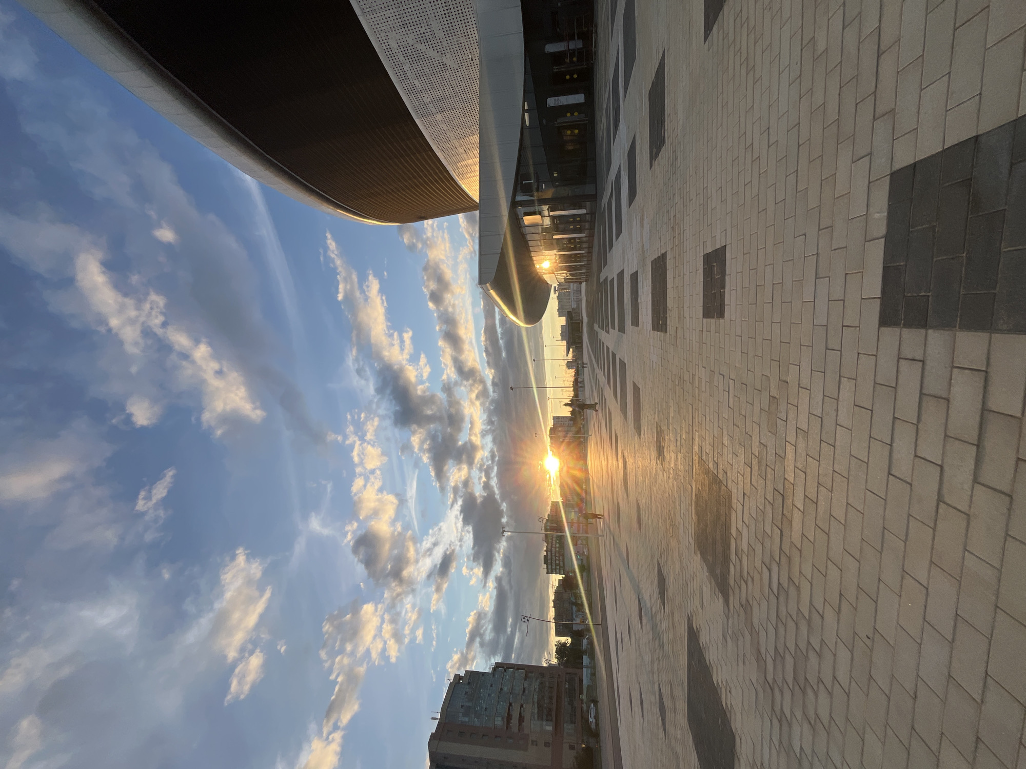 The image depicts an outdoor scene during sunset in a modern urban environment. The sky is a mix of blue and white clouds, illuminated by the setting sun which is prominently visible in the center left of the frame, casting a warm glow and lens flares across the scene. In the foreground, a wide paved area with a patterned brickwork surface stretches into the distance. This area is marked by alternating light and dark rectangular tiles embedded within the brickwork. To the right, a large, curved modern building with a dark upper section and a lighter, perforated facade occupies a significant portion of the frame. The lower level of this building features a glass entrance with visible doors. To the left, several multi-story buildings are visible, characteristic of a city skyline. Streetlights are spaced along the paved area. In the middle ground, there are a few figures, one walking and another on a scooter, suggesting public activity. The overall impression is a contemporary cityscape bathed in the warm light of sunset.
Based on visual cues such as the architectural style of the buildings and the urban planning, this location strongly resembles the area around the Nizhny Novgorod Stadium in Nizhny Novgorod, Russia. The stadium's distinctive curved architecture and the surrounding modern paved public space are consistent with the design of the area around major sports venues.
