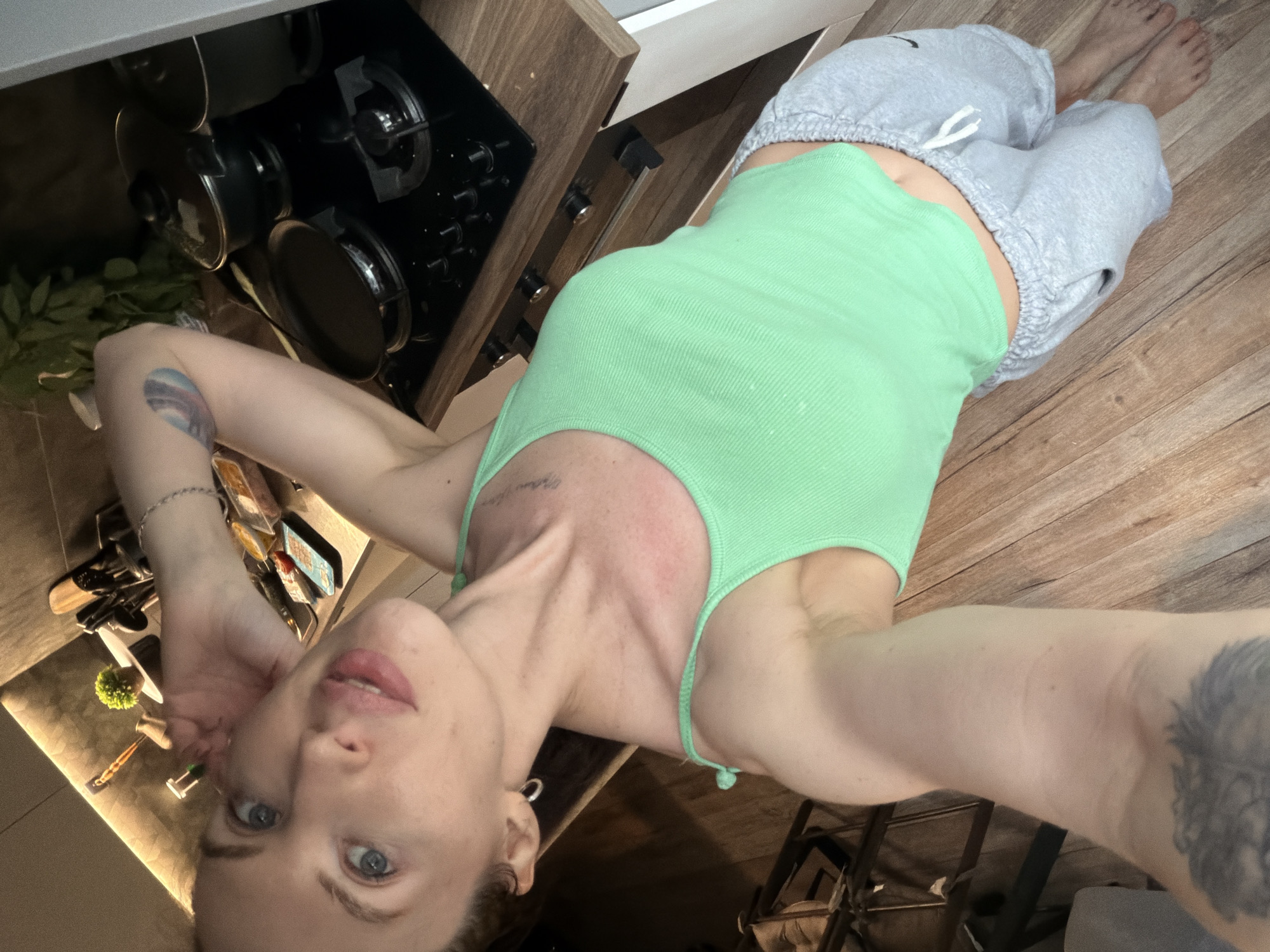 A woman wearing a green tank top and gray sweatpants is lying on a wooden floor. She is positioned near a kitchen counter with a stovetop and drawers. Various utensils, dishes, and greenery are visible on the counter. The location is likely an interior domestic space. There is no information in the image that indicates a location in Perm, Russia.