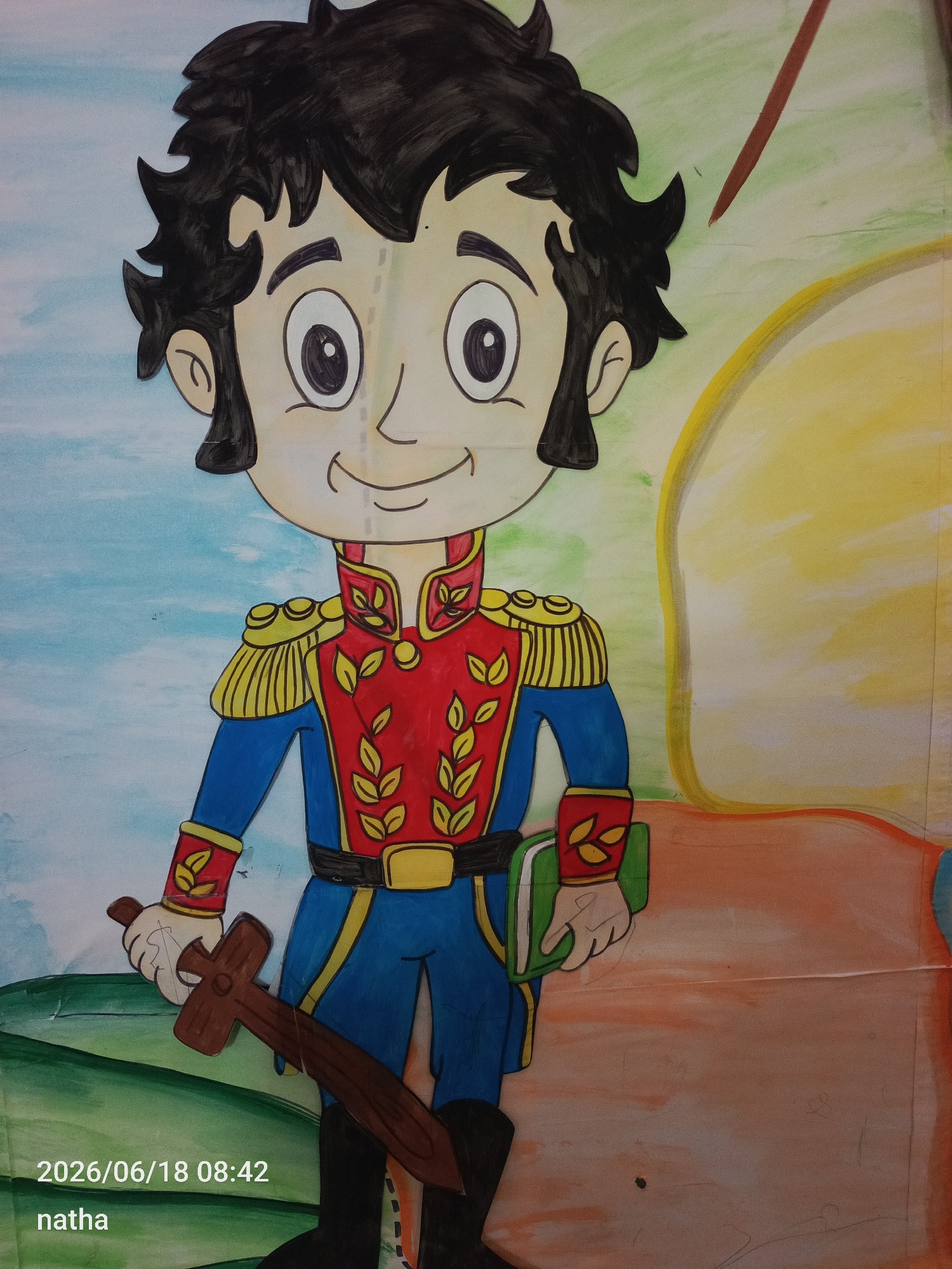 This is a photograph of a colorful, cartoon-style drawing of a young boy dressed as a soldier or a historical figure. The drawing is made on paper and appears to be a cut-out or part of a larger mural.
The boy has black, spiky hair, large expressive eyes, and a cheerful smile. He is wearing a red jacket with gold trim and epaulets, blue pants, and black boots. The jacket is decorated with yellow leaf-like motifs. He is holding a wooden sword in his left hand and a green book in his right. The background of the drawing is painted with broad strokes of blue, green, yellow, and orange, suggesting a landscape or abstract scene. The style is reminiscent of children's book illustrations.
Visible text includes a date and time stamp in the lower left corner: "2026/06/18 08:42" and the name "natha".
The image is taken in San Sebastián, Venezuela. While the drawing itself doesn't offer specific location cues, the presence of the text might indicate an event or artwork created for a school or community event in that area. The emotion conveyed by the drawing is one of joy and possibly heroism or learning, given the sword and book. The time stamp suggests it was captured in the morning.