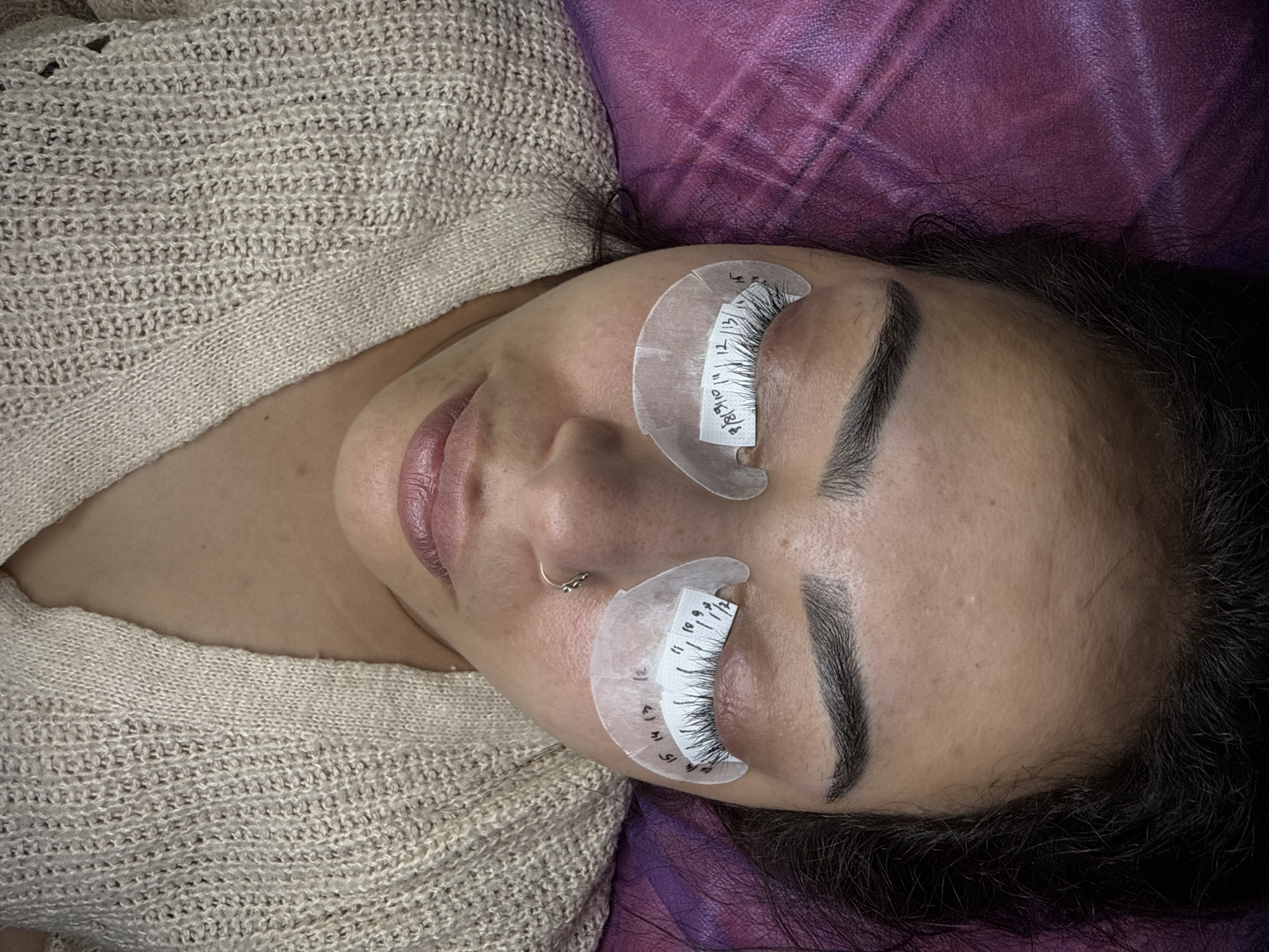 A close-up, overhead shot captures a person lying down with their eyes closed and undergoing a lash lift procedure. The individual is wearing a beige knit sweater and rests on a purple surface. Their eyelashes are separated by white under-eye pads, which are marked with black numbers indicating lash lengths. The lighting is bright and even, illuminating the subject's face and the details of the procedure. The overall scene suggests a beauty treatment taking place in a controlled environment. There is no indication of the city of Pamplona, Spain.