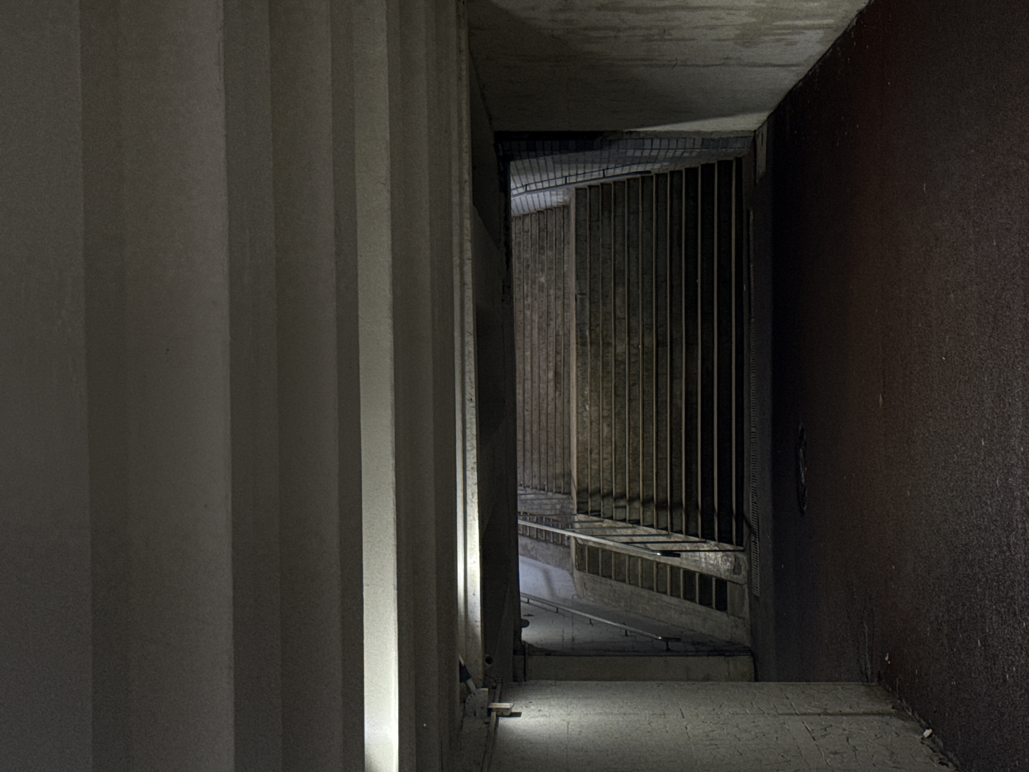 The image depicts a concrete structure, likely an underpass or tunnel, located in Perm, Russia. Vertical concrete pillars are visible on the left. A metal grating enclosure with vertical bars extends along the center of the image, obscuring a downward-sloping ramp or stairway. The right side is a dark, textured wall. The scene is illuminated by artificial light, casting shadows and highlighting textures. No people are visible. The overall atmosphere is dimly lit and industrial.