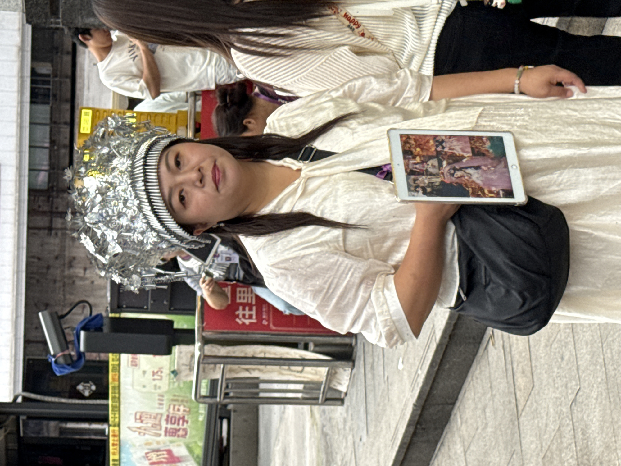 Here is a detailed description of the image:
A medium shot shows a woman holding up a tablet, standing outside on a paved surface. The woman has a silver headpiece and pigtails, and she is wearing a light-colored blouse and a black shoulder bag. The tablet is showing an image of what appears to be artwork or a digitally altered image. The background includes other people, including someone wearing a white shirt, and what appears to be a bus stop or a public transit sign with Chinese characters. Based on the sign and the environment, it is likely that the location is Yuzhong District, China.