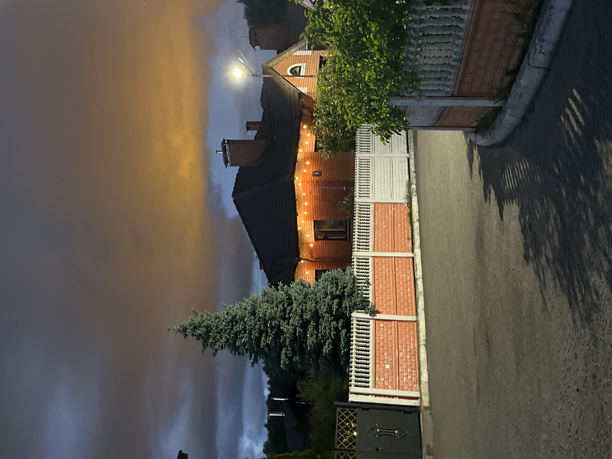 This media file captures a nighttime scene in a residential area of Ореховка, Russia. The central subject is a two-story house constructed from reddish-orange brick, featuring a dark, possibly tiled roof and multiple chimneys. String lights (fairy lights) illuminate the facade of the house, adding a warm glow. A bright street light mounted on a pole provides additional illumination to the scene.
To the left of the house, a tall, dense evergreen tree, likely a spruce or fir, stands prominently. In the foreground, a fence made of reddish-orange brick and white metal sections borders the property, with a dark, ornate metal gate visible at the bottom. A paved road or driveway runs parallel to the fence.
The most striking detail is the sky, which exhibits a dramatic color variation. On the left side, it appears a deep, dark grey-blue, transitioning towards the right into a distinct orange-yellow hue, suggesting either significant light pollution reflecting off dense cloud cover or a unique atmospheric event. No people or specific activities are visible, giving the scene a quiet and still ambiance. There is no visible text in the image.