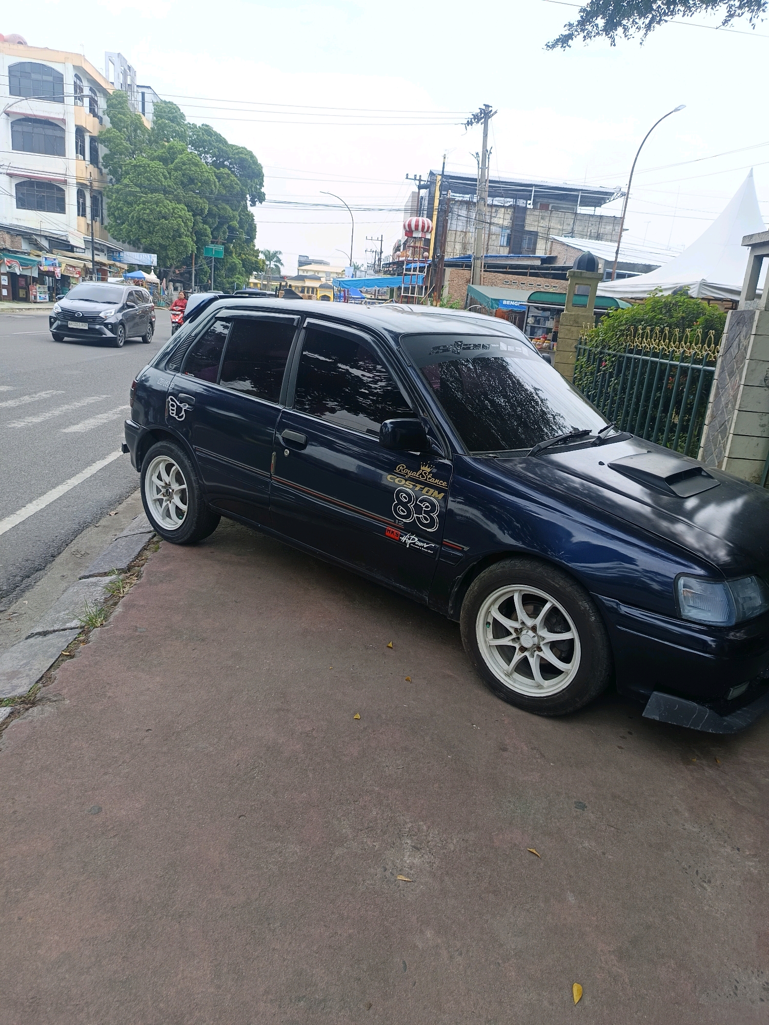 A dark blue hatchback car is parked on the side of a street in Medan, Indonesia. The car has white aftermarket rims and several stickers on its body, including one with "Royal Stance Custom 83" and another with a hand gesture. A black hood scoop is visible on the car's hood. In the background, there are buildings, trees, and other vehicles on the street, including a gray sedan. The street appears to be a busy urban area.