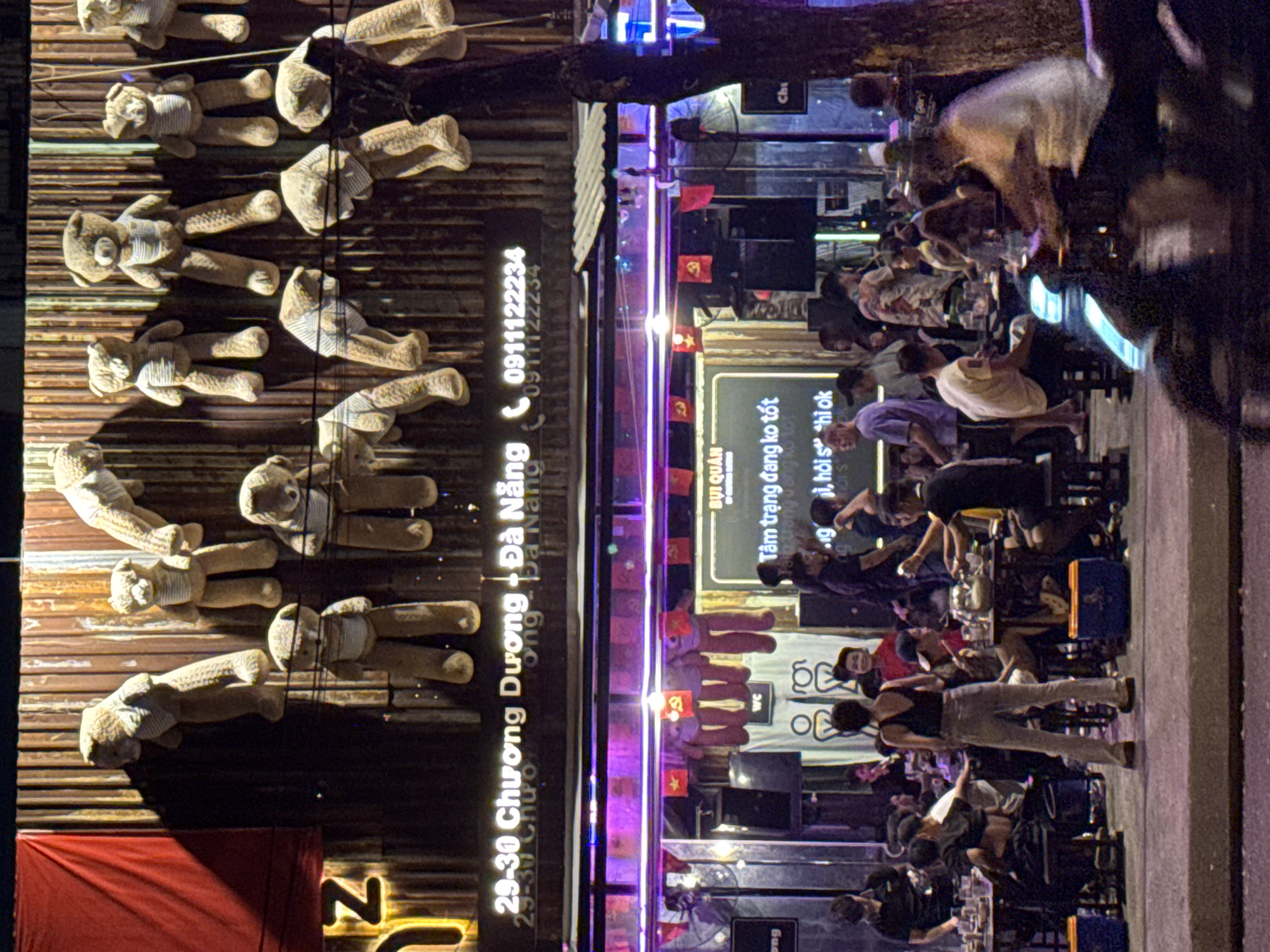 The image depicts an outdoor scene at night, likely a street-side establishment in Da Nang, Vietnam, identified by the signage "29-30 Chuong Duong + Da Nang" and the phone number "0911122234". The establishment appears to be a bar or restaurant, with tables and people seated and standing around. The exterior features a corrugated metal wall decorated with numerous teddy bear figures hanging at various levels. Some of the teddy bears are worn and weathered. In the foreground, a street or sidewalk is visible, with a person on a motorbike moving from right to left, causing motion blur.
Above the seating area, there is a sign with Vietnamese text that includes "BUI QUAN" and "Tam trang dang ko tot," which roughly translates to "Bui Quan" and "Mood not good." There are also flags with red and yellow colors displayed, some of which appear to be communist flags with hammers and sickles. Neon lights and string lights provide illumination, casting a purple hue over parts of the scene.
The people in the image are dressed casually and appear to be socializing and dining. The overall atmosphere suggests a lively, informal gathering place. The background shows the dark facade of the building and the night sky. The image captures a slice of urban nightlife in Da Nang.