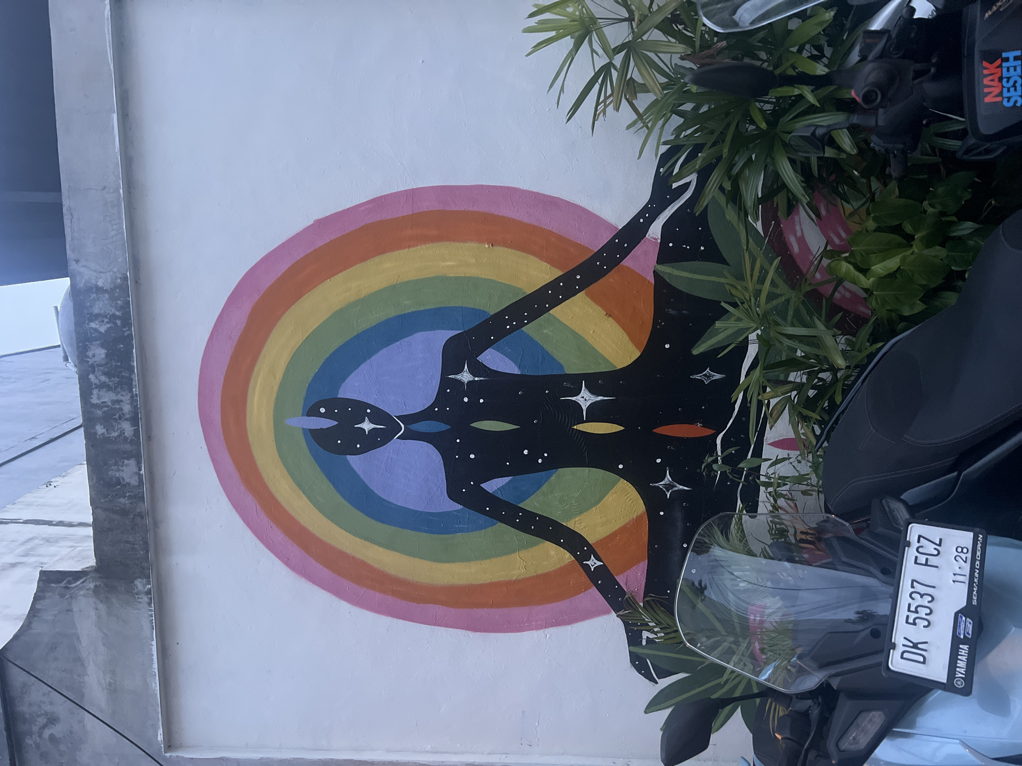 A mural is painted on a white wall, featuring a figure outlined in black with a star pattern, superimposed over a rainbow circle. The colors of the rainbow are, from the outside in, pink, orange, yellow, green, blue, and purple. The mural is partially obscured by green plants and a parked motorbike with a license plate reading "DK 5537 FCZ". The license plate also has "Yamaha" written on it. The photograph was taken in the city of Tibubeneng, Indonesia.