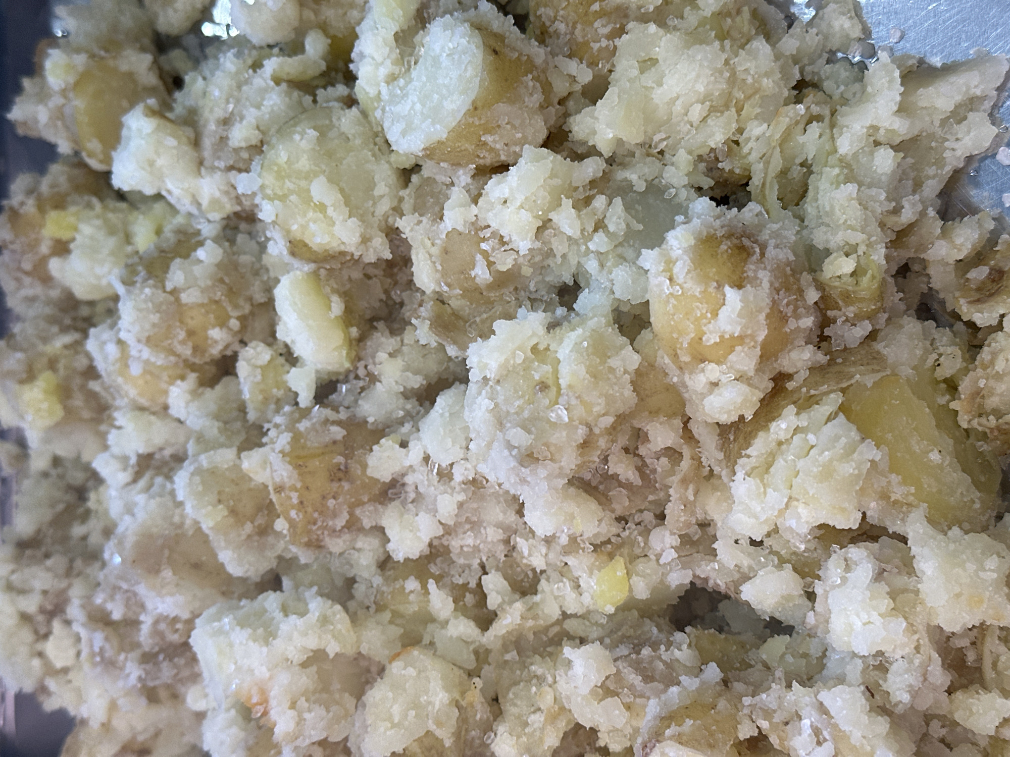 The image is a close-up, overhead shot of a pile of cooked, mashed potatoes in a metal tray. The potatoes are pale yellow and white, with a lumpy, uneven texture. Some pieces show remnants of potato skin, which is brown and slightly rough. The potatoes appear to be seasoned with coarse salt, which is visible as small, white crystals scattered throughout the dish. The lighting is bright and even, casting minimal shadows. There are no people or other objects visible in the frame, except for the metal tray, which appears to be silver or stainless steel and has a slightly reflective surface. The image is a still life of food preparation.