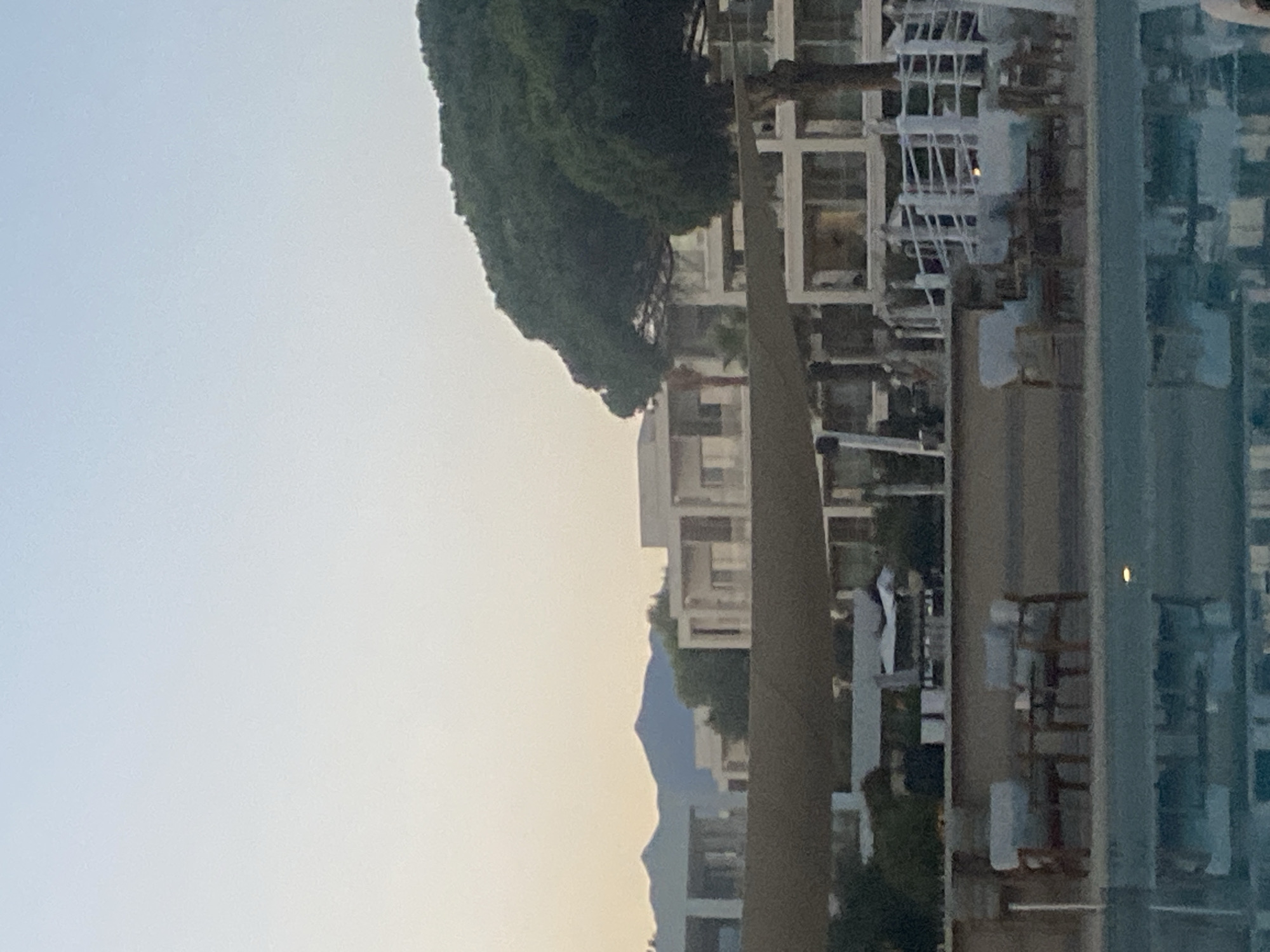 This is a vertically oriented, out-of-focus, medium shot photograph taken from a high vantage point, possibly a balcony or window. The scene depicts a modern resort or apartment complex with white buildings, glass balconies, and an abundance of lush green trees. In the distance, a mountain range is visible, silhouetted against a hazy sky, suggesting a clear day. The water in the foreground is reflective, mirroring the buildings and trees, and the presence of beach chairs and umbrellas implies a coastal location. The overall impression is one of a tranquil, upscale vacation destination, likely during daylight hours with fair weather. The image is characterized by soft lighting and a dreamy, almost abstract quality due to its blurriness. There are no people or discernible activities visible, emphasizing the quiet and serene atmosphere of the resort. The location context of Estepona, Spain, aligns with the visual cues of a coastal resort town with mountains nearby.