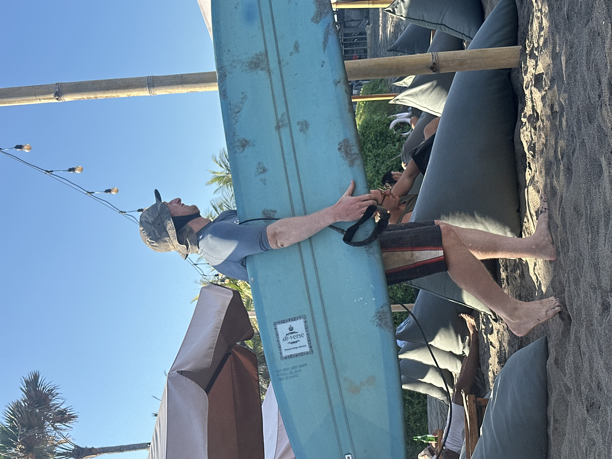 In North Kuta, Indonesia, on a beach, a man wearing a camouflage hat and a blue rashguard walks across the sand carrying a light blue surfboard. He is wearing dark shorts with red and white stripes. The surfboard has a sticker on it with the text "de.verse" and "Modern Surf Creations". In the background, there are several large, dark gray beanbag chairs on the sand, and beyond that, some vegetation and structures. String lights with Edison bulbs hang from a bamboo pole. A palm tree and a section of an umbrella are visible in the upper left. The sky is clear and blue.