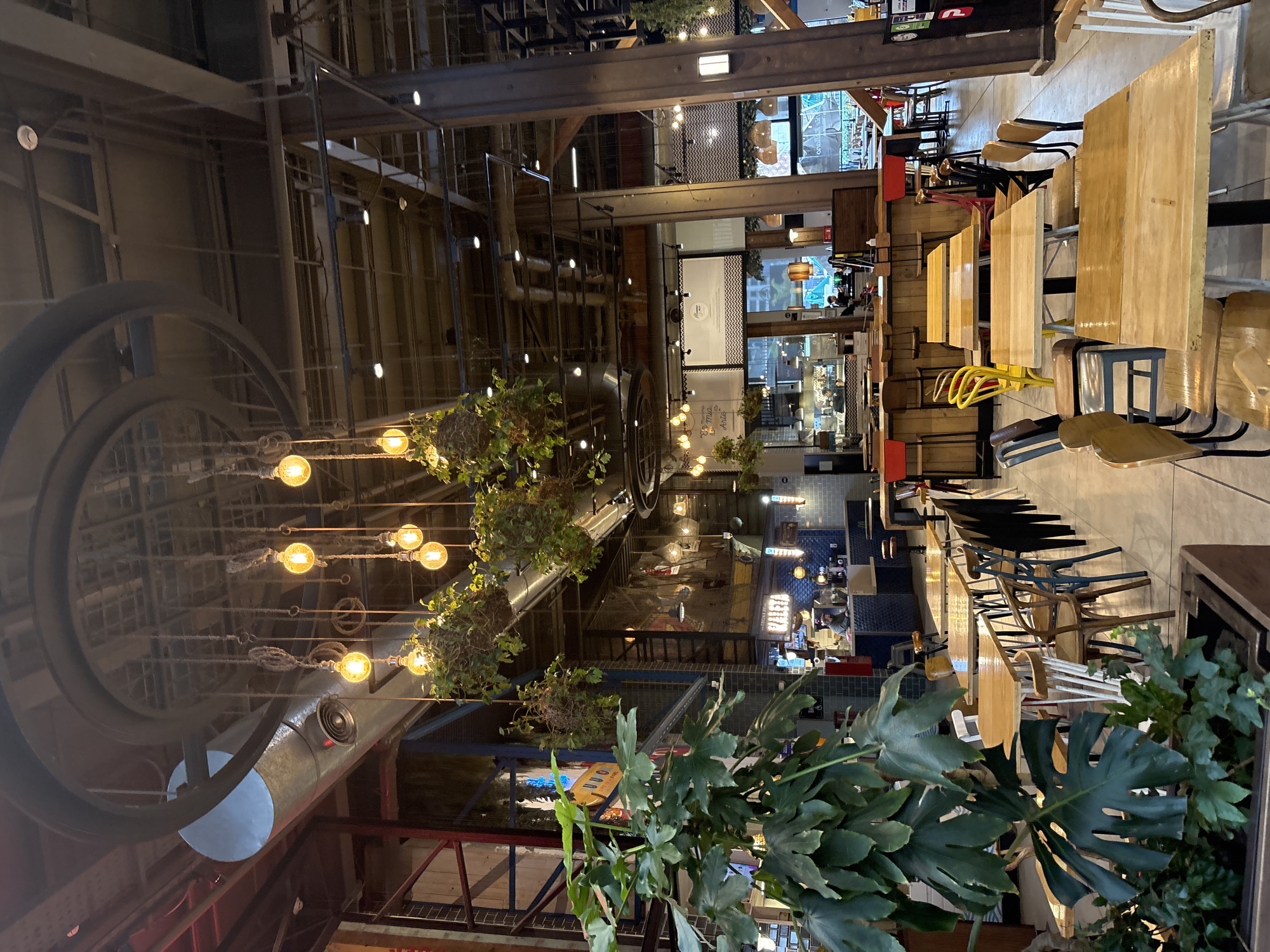 The image depicts the interior of a bustling food market in Santiago, Chile. The scene is characterized by a rustic-industrial aesthetic, with exposed beams, metal ductwork, and Edison-style lightbulbs hanging from the ceiling. Lush green plants are strategically placed throughout the space, some suspended from above in hanging baskets, adding a touch of nature to the urban environment.
Numerous tables and chairs, made of wood and metal, are arranged to accommodate diners. Some tables are set for immediate use, while others appear to be waiting for customers. Behind the tables, a vibrant food counter area is visible, with staff members attending to orders and food preparation. The counters are lined with blue tiles and adorned with various food service equipment.
The overall atmosphere is lively and inviting, suggesting a popular dining destination within the city. The architecture and decor imply a contemporary design that blends industrial elements with natural touches, creating a unique and engaging space for patrons to enjoy their meals. The presence of signage in Spanish further confirms the location in Chile.