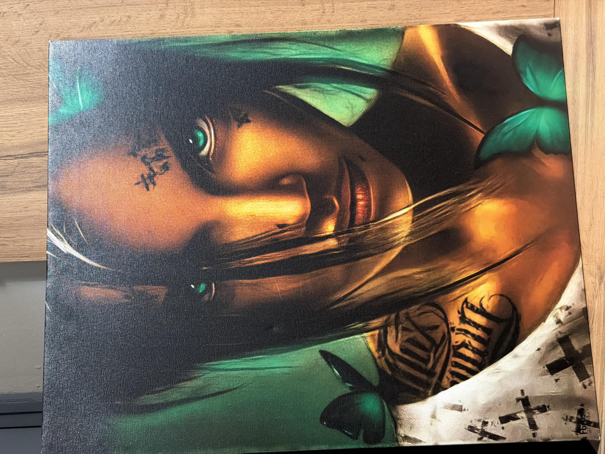 The media file shows a canvas print of an artwork. The artwork is a portrait of a person with striking green eyes and a tanned complexion. The person's face is angled, with strands of dark hair obscuring parts of it. There are markings on the forehead. The artwork features elements of fantasy or surrealism, with green butterfly-like shapes adorning the upper right and lower right portions of the canvas. The background is a mix of dark and emerald green tones, with hints of golden light illuminating the person's skin. Visible on the person's arm is a tattoo with bold black lines. The lower right corner of the canvas has a white area with black cross-like markings and a small white rectangle with dark text. The canvas is placed on a wooden surface, suggesting it is being displayed or stored. The lighting is natural and comes from above. No specific time of day or weather is indicated. The emotions conveyed by the portrait are enigmatic and intense due to the direct gaze and stylized presentation.