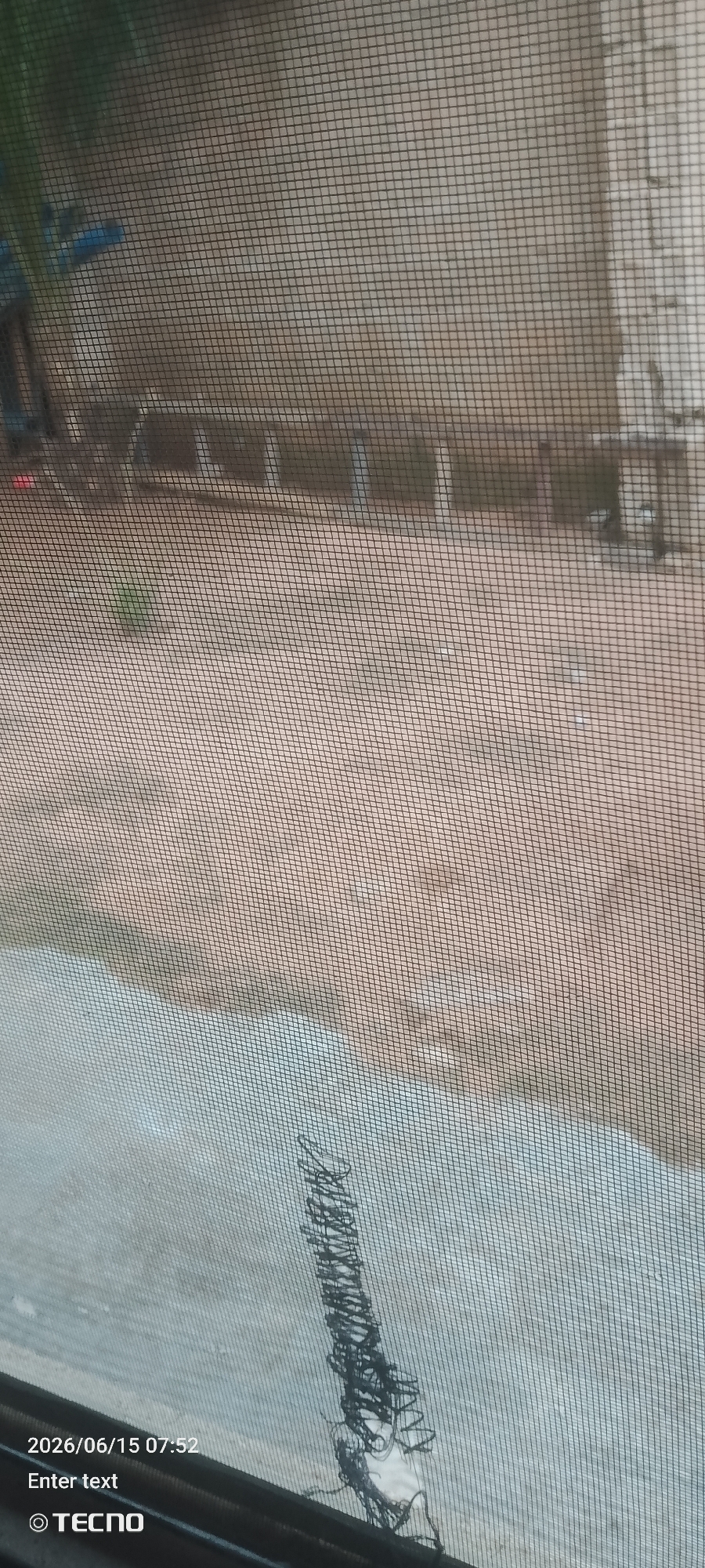 The image shows a view through a window screen. The scene outside appears to be a dirt or gravel area with some structures in the background that are out of focus. There is a hint of greenery on the left side of the image. In the lower right corner of the image, there is a dark, tangled object that appears to be a piece of wire or string. The date and time "2026/06/15 07:52" are visible in the lower left corner, along with the text "Enter text" and a TECNO logo.
