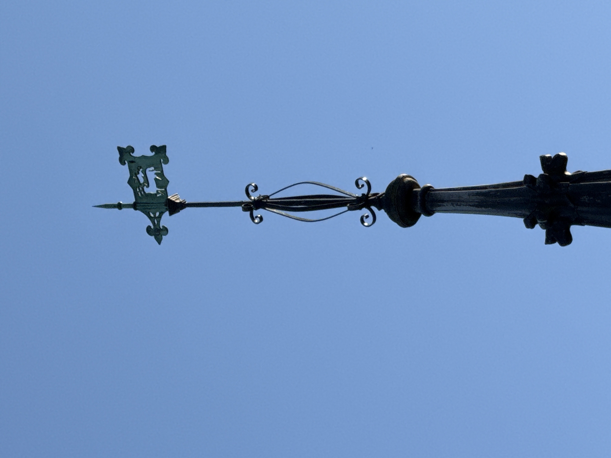 The image shows a weather vane against a clear blue sky. The weather vane is made of dark metal and is elaborately designed with a decorative crest on one end. The overall design is intricate, with a series of stylized curls and scrolls. The location is not specified, and the prompt only gives a general location as Dacorum, United Kingdom. Therefore, the image details are limited to the visible weather vane and sky.