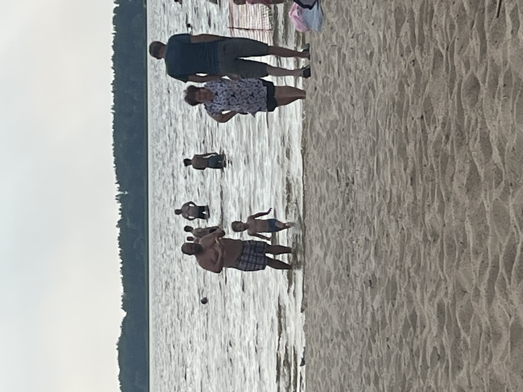 The image shows a group of people at a beach, with the water and sand in the foreground. The setting appears to be a lake or calm body of water with a tree-lined shore in the background. The time of day seems to be overcast, possibly late afternoon or evening, as the light is muted.
In the water, several individuals are visible. Towards the left, a man in plaid shorts is holding a child, and another child is standing nearby. Further back, there are other individuals in the water, some appearing to be adults and others possibly children. On the sandy shore, a man in a dark t-shirt and shorts stands next to a woman wearing a patterned top and shorts. A pink bag and a white object, possibly another bag or clothing item, are also on the sand behind them.
The people seem relaxed and engaged in typical beach activities, such as wading in the water or relaxing on the shore. The overall mood appears to be casual and recreational. There is no visible text in the image. The location context is described as Загародскі сельскі Савет, Belarus.