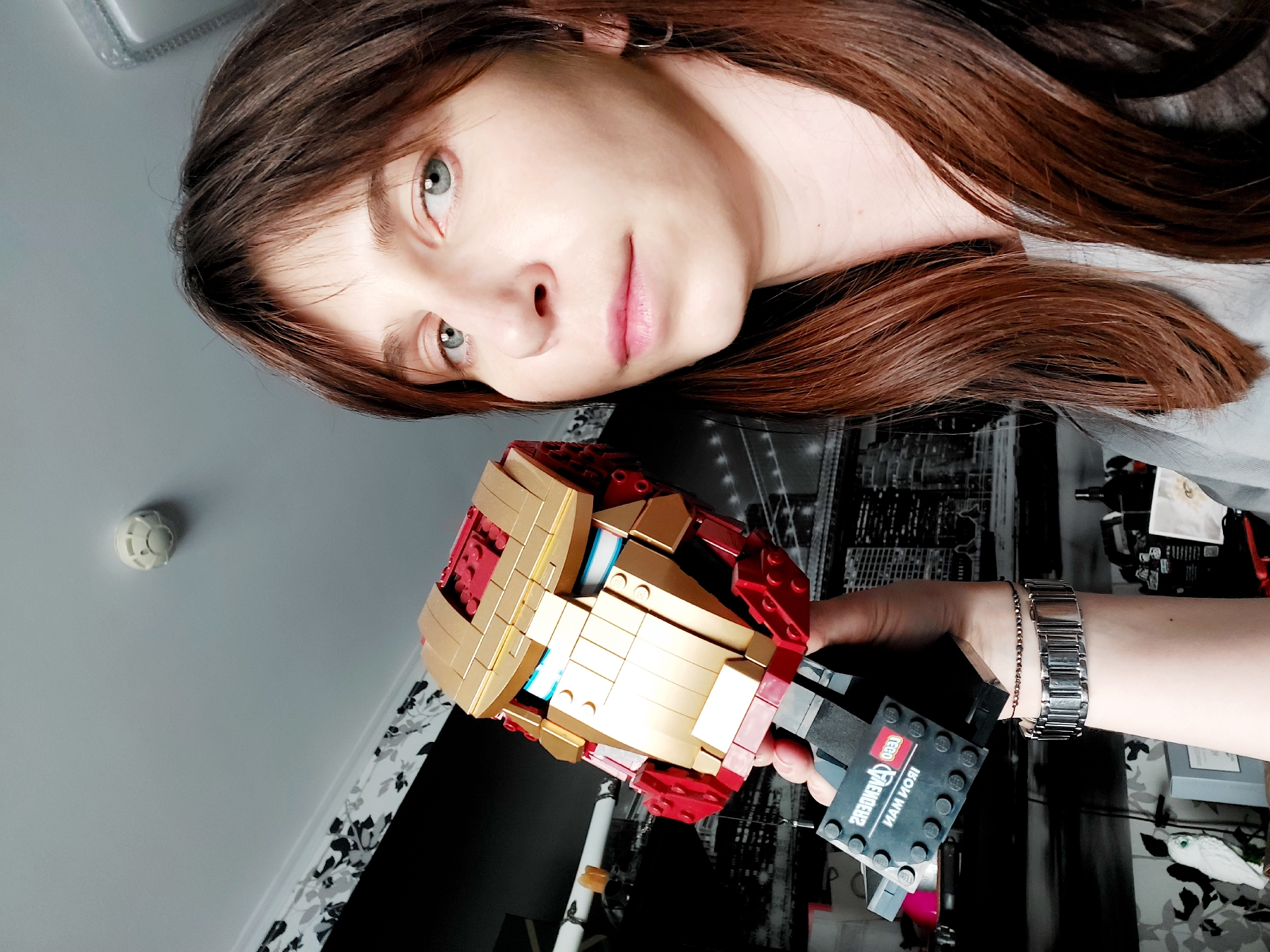 A young woman with light skin, light eyes, and brown hair is holding a Lego Iron Man mask. She is wearing a silver watch on her left wrist. The background shows a room with a black and white patterned wall. A smoke detector is visible on the ceiling. It is not possible to discern the location of the photo as Izhevsk, Russia from the image.