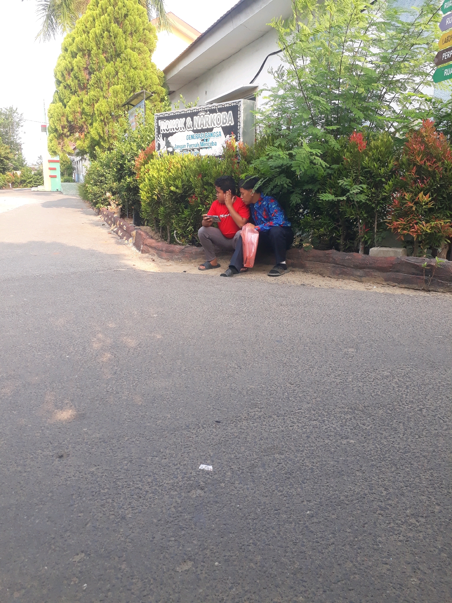 In Pangkalpinang, Indonesia, two young males are seated on a log near a lush green hedge. The male on the left, wearing a red shirt and gray pants, holds a mobile phone and appears to be looking at it. The male on the right, dressed in a blue shirt with a patterned design and dark pants, is seated next to him, holding a red plastic bag. Behind them, a sign reads "ROKOK & NARKOBA GENERASI BANGSA". The foreground features an asphalt road, with a small crumpled piece of paper visible on its surface. The background reveals a building with a white exterior and a sloped roof, bordered by trees and dense foliage.