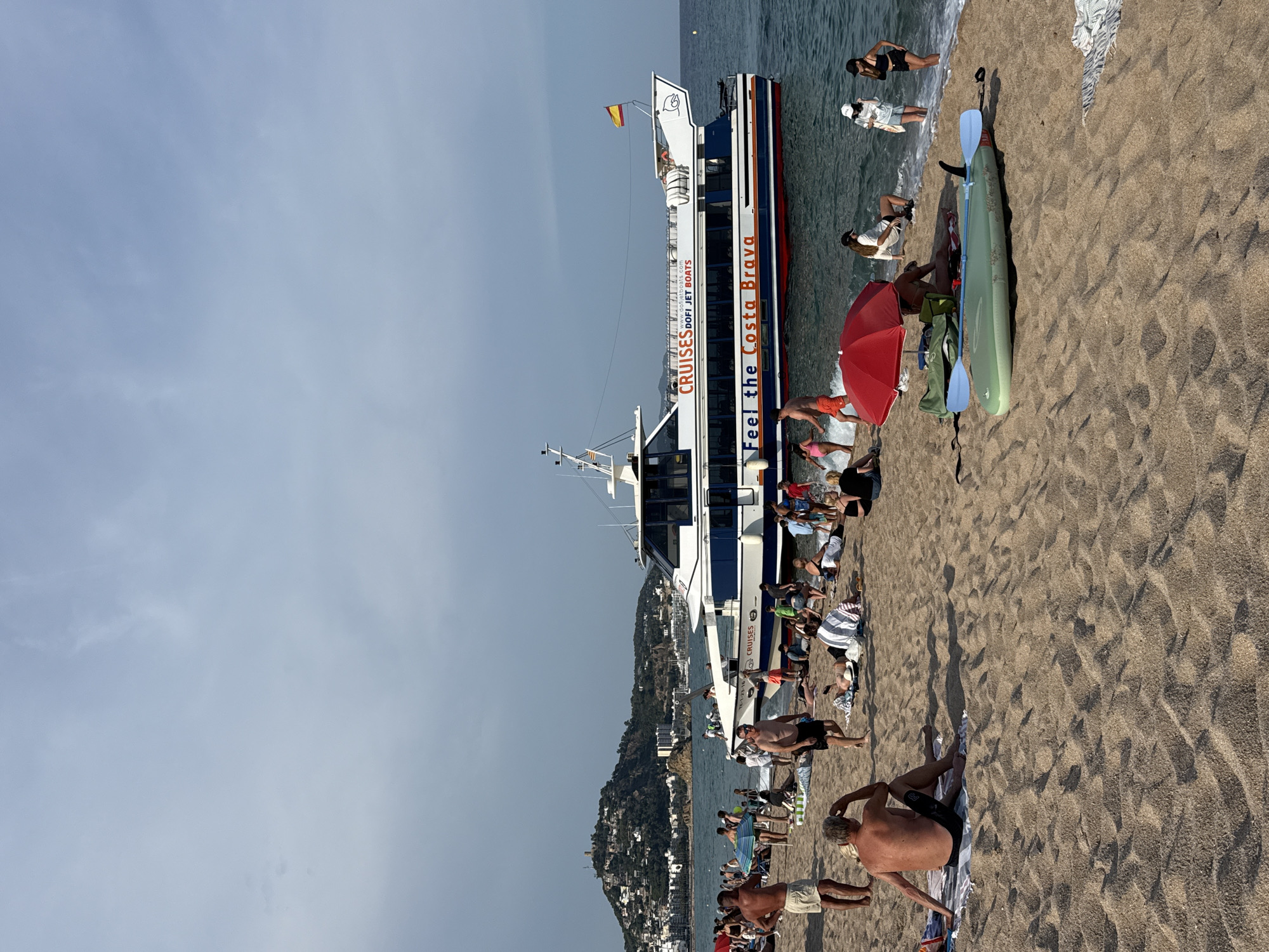 This media file depicts a sunny day at a crowded beach in Blanes, Spain. The scene is dominated by a large white and blue boat docked near the shore, with "Feel the Costa Brava" and "CRUISES Dofi JET BOATS" visible on its side, along with a website address. A Spanish flag flies from the boat's mast.
On the sandy beach, numerous people are engaged in various leisure activities. Some are sunbathing, sitting on towels or beach chairs, while others are wading in the shallow, wavy water. A bright red umbrella provides shade for a group of beachgoers. A green kayak with two paddles lies on the sand, suggesting recreational water activities.
The time of day appears to be afternoon, given the strong sunlight and long shadows cast on the sand. The weather is clear and bright, with a few wispy clouds in the sky. In the background, a green, hilly coastline covered with buildings slopes down towards the sea, indicating a developed coastal area. The overall mood seems relaxed and festive, with many people enjoying the summer day.