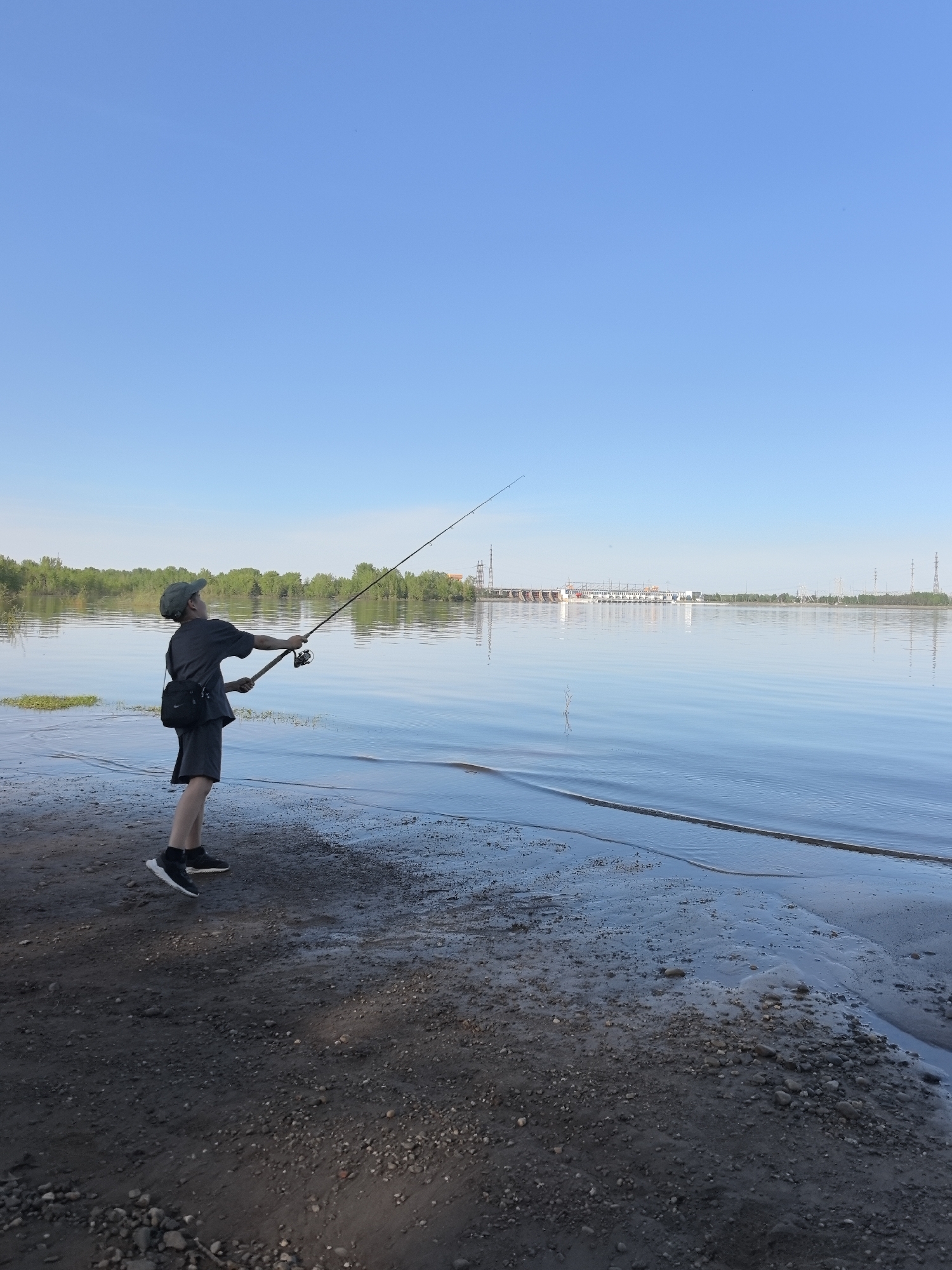 A young person is fishing from the edge of a body of water, likely a river or large lake. The person is standing on a dark, wet sandy or muddy bank, holding a fishing rod and looking upwards toward the end of the rod. They are wearing dark clothing and shoes, with a cap and a bag slung across their shoulder.
In the background, across the water, there is a landscape including trees and what appears to be a hydroelectric dam or other industrial structure. Several electrical towers are visible in the distance. The sky is clear and blue. The overall impression is that of a calm, sunny day at a waterfront location.
The location of Noviy, Russia is included. There is no way to verify this from the photo.