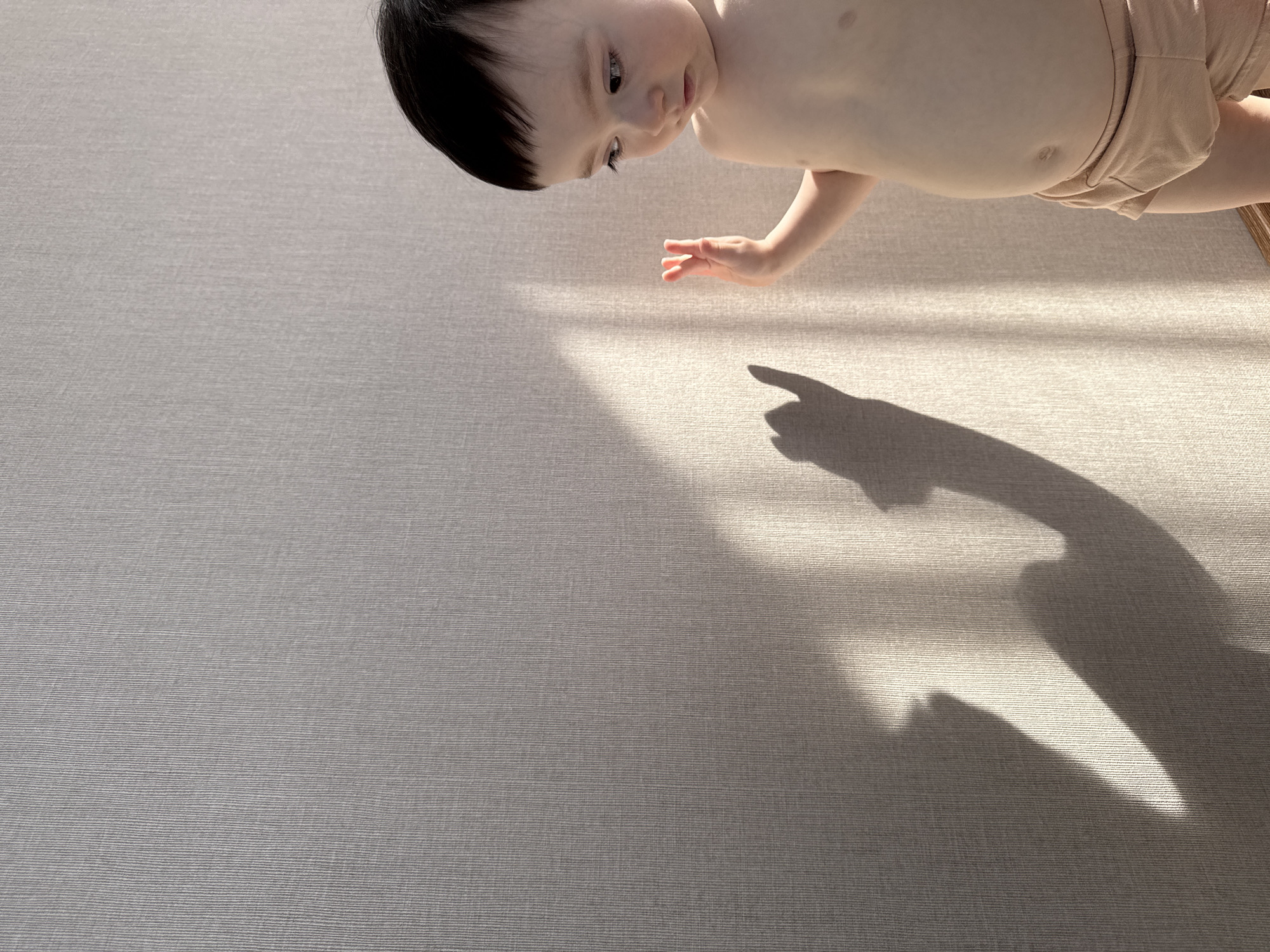 The scene depicts an indoor environment, presumably in Izhevsk, Russia. A single infant, appearing to be between 6 and 18 months of age, with dark hair, is positioned on the right side of the frame, facing left. The infant is shirtless, wearing light-colored, possibly beige or cream-colored, undergarments resembling shorts. Its head is turned slightly to the left, with eyes gazing downward. The infant's right arm is raised, with fingers slightly splayed. The background consists of a light-colored wall, possibly beige or off-white, featuring a consistent vertical linear texture. Direct sunlight enters the scene from the right, illuminating the infant's right side and casting pronounced, elongated shadows on the wall. A distinct shadow of the infant's raised right hand and arm is visible on the wall, to the left of its actual hand. Other shadows, including what appears to be part of the infant's body, are cast on the lower left portion of the wall. A narrow strip of a darker wooden baseboard is visible at the very bottom right corner of the frame. The infant appears to be observing or interacting with the interplay of light and shadow on the wall.