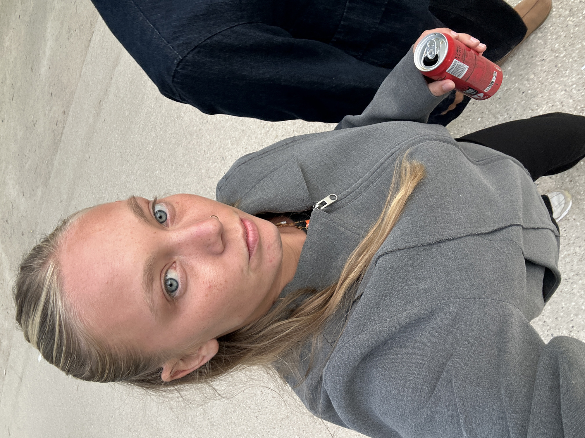 The image shows a fair-skinned person with light blonde hair and blue eyes, wearing a gray zip-up hoodie. The person is holding an open can of Coca-Cola Zero. A dark blue denim-clad leg and a person wearing tan shoes are visible in the background, along with another leg in black pants and a white shoe. The location is most likely indoors with a light-colored floor. There is no information that would place the image in the city of Paris, France.