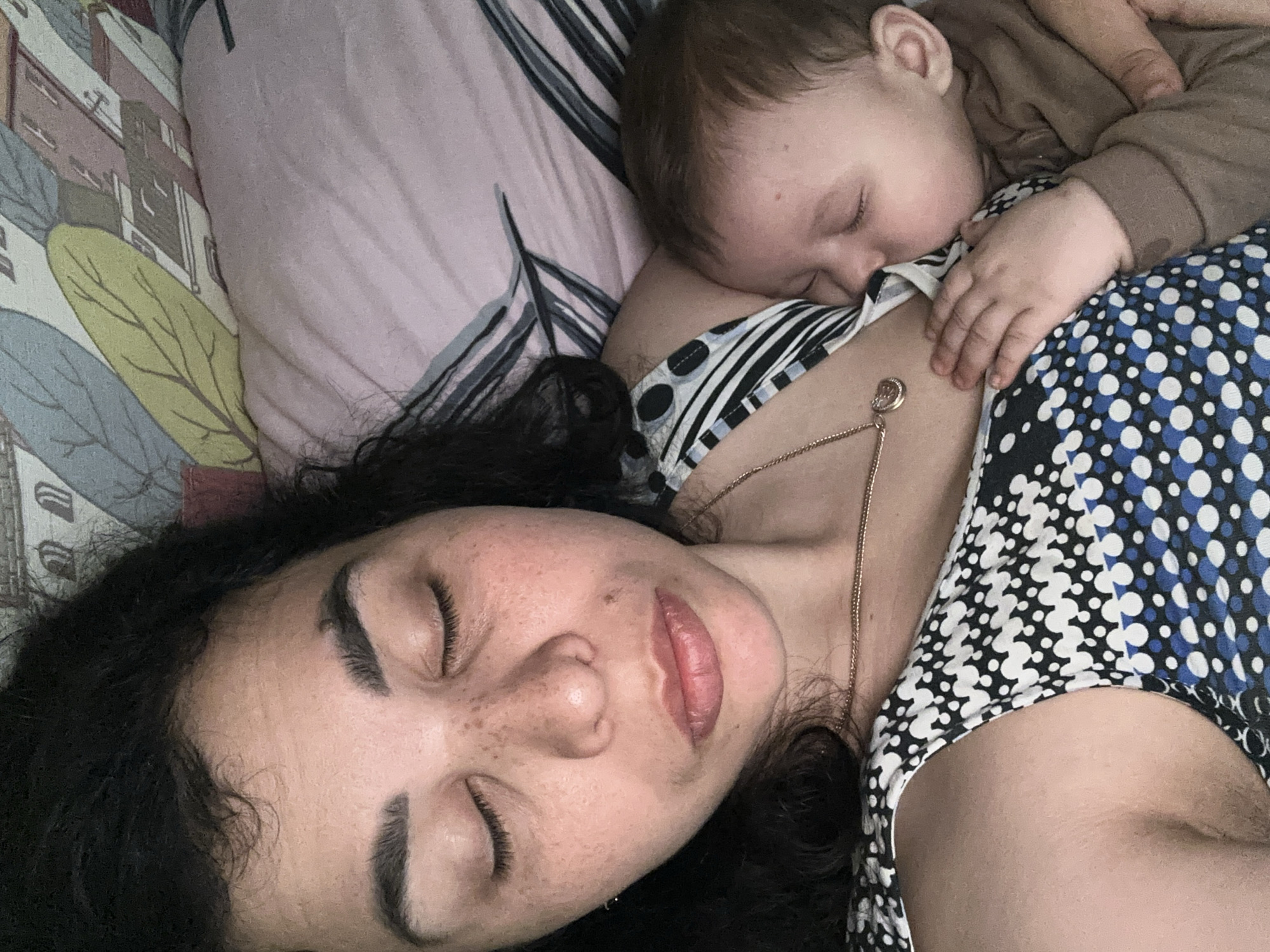 An overhead close-up shot captures a woman and a baby sleeping peacefully together on a bed. The woman, lying on her back, has her eyes closed, displaying long, dark, curly hair, visible eyelashes, well-defined eyebrows, and freckles across her nose and cheeks. Her lips are slightly parted. She wears a patterned top featuring black, white, and blue polka dots or geometric shapes, and a delicate gold chain necklace with a small, circular pendant rests on her chest.
The baby is nestled on the woman's shoulder and chest, also asleep with closed eyes. It wears a light brown long-sleeved top, and one tiny hand rests gently on the woman's chest.
The setting is indoors, presumably a bedroom. A light pink pillow with subtle darker patterns is visible behind the woman's head. The bedding next to her features a distinctive pattern of stylized buildings and large, muted green and grey leaves against a lighter background. The image conveys a sense of deep serenity, comfort, and a close, tender bond between the two subjects. There is no visible text in the image.