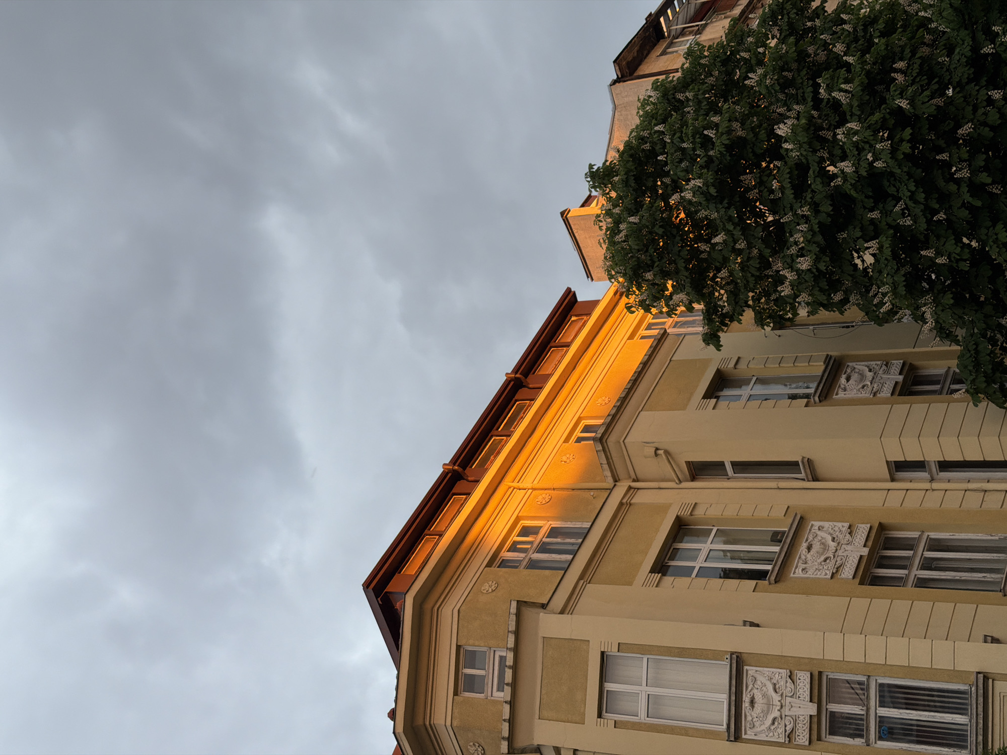 Captured from a low angle, the image showcases a segment of a multi-story building against a backdrop of a cloudy sky. The building's facade is painted in warm yellow tones, with notable architectural details such as decorative reliefs beneath some windows. A tree with lush green foliage partially obscures a portion of the building. The lighting suggests either sunrise or sunset, casting a golden glow on the building's edge. The location is identifiable as Sofia, Bulgaria, by the building style and architectural features.