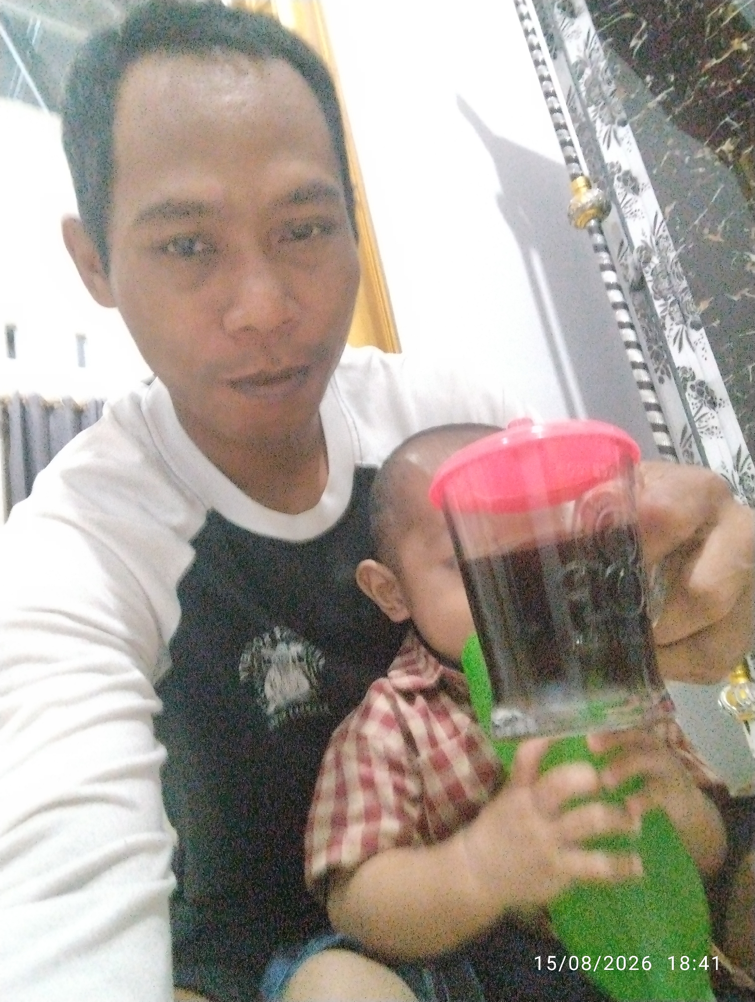In this close-up, selfie-style photograph, a man with dark skin and short hair is prominently featured, looking directly at the camera. He is wearing a white t-shirt with black sleeves and a black graphic design on the chest. To his right, a baby is partially visible, also looking away from the camera. The baby is wearing a red and white checkered shirt. In the foreground, a clear plastic cup with a pink lid holds a dark liquid and a green object, possibly a straw or a holder. The background is blurred, showing hints of a wall with a decorative pattern and vertical blinds. A timestamp in the lower right corner reads "15/08/2026 18:41". The lighting suggests an indoor environment, possibly a home. The man's left hand is holding the cup, and the baby's hands are visible around the green object. The overall impression is a candid moment captured between the man and the baby.