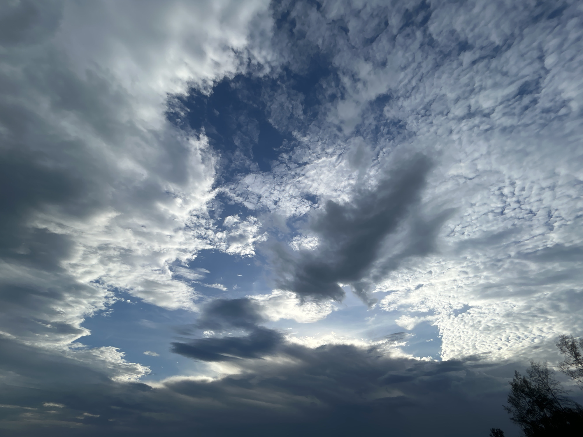 A dramatic sky over Koh Phangan, Thailand, features a dense array of clouds. The clouds range from dark, heavy storm clouds at the bottom of the frame to lighter, wispy clouds higher up. A central opening in the clouds reveals a patch of bright blue sky, with sunlight breaking through and illuminating some of the cloud edges. The overall impression is one of dynamic weather conditions. The lower edge of the image shows a dark silhouette of vegetation, possibly trees, against the lighter sky, suggesting an outdoor, possibly coastal, location.