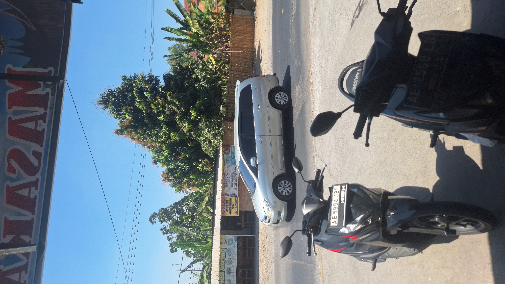 In the city of Widodaren, Indonesia, under a bright, clear blue sky, a silver Toyota Innova minivan is captured in motion on a paved road. The minivan is traveling from left to right. In the foreground, two parked motorcycles are visible, one black with a license plate reading "AE 5994 JBF," and another partially obscured motorcycle. Behind the road, a row of buildings with corrugated metal roofs is present. These buildings appear to be small businesses, with signage indicating food offerings like "Nasi Goreng" (fried rice) and "Soto Ayam Kampung" (kampung chicken soup). Lush green trees, including banana trees and other leafy vegetation, are abundant, with one particularly large, bushy tree dominating the middle ground. A bamboo fence runs along the edge of the property behind the trees. Above the scene, a sign with red lettering reads "MASAKAN NASI GORENG."
