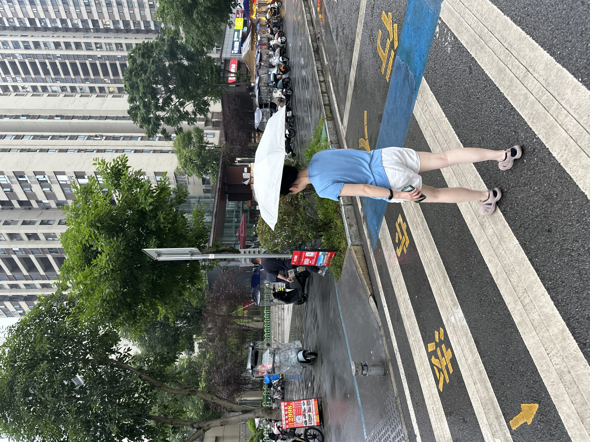 In the city of Chongqing High-tech Zone, China, a woman with dark hair, wearing a blue t-shirt and white shorts, walks across a wet pedestrian crossing, holding a white umbrella over her head. She is wearing pink sandals and has a smart watch on her left wrist. The pedestrian crossing has white and yellow markings on the asphalt. In the background, several parked electric scooters are visible, some with protective covers. A man in a black t-shirt and dark pants is also present, walking away from the camera. Further back, tall apartment buildings dominate the skyline, with trees and greenery interspersed between them. The street is wet, indicating recent rain. Red promotional signs are also visible near the parked scooters.