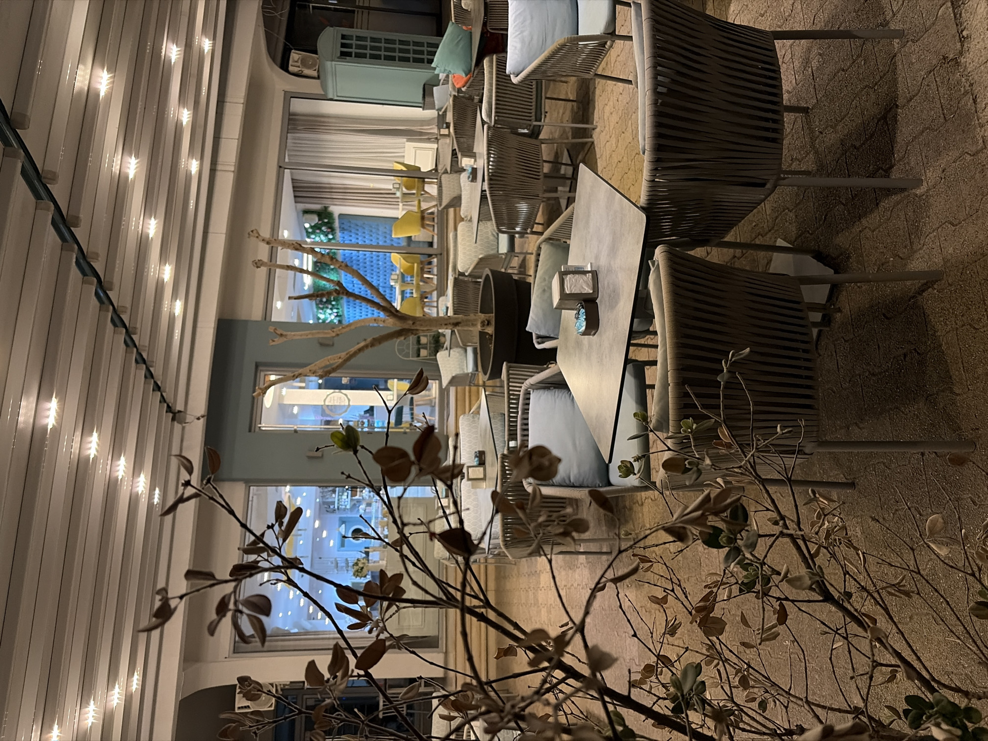 The image depicts an outdoor seating area of a restaurant or cafe, likely in the city of Bar, Montenegro, given the context provided. The scene is set in the evening, indicated by the soft lighting.
Several tables with modern, woven chairs are arranged in a row. The tables are made of light-colored material. Each table has a napkin holder.
The backdrop consists of building facades with large windows showing brightly lit interior spaces. There are some decorative elements, including light fixtures. A decorative plant stands in the foreground.
The overall atmosphere is inviting and relaxed, suggesting a popular spot for socializing and dining in Bar, Montenegro.
