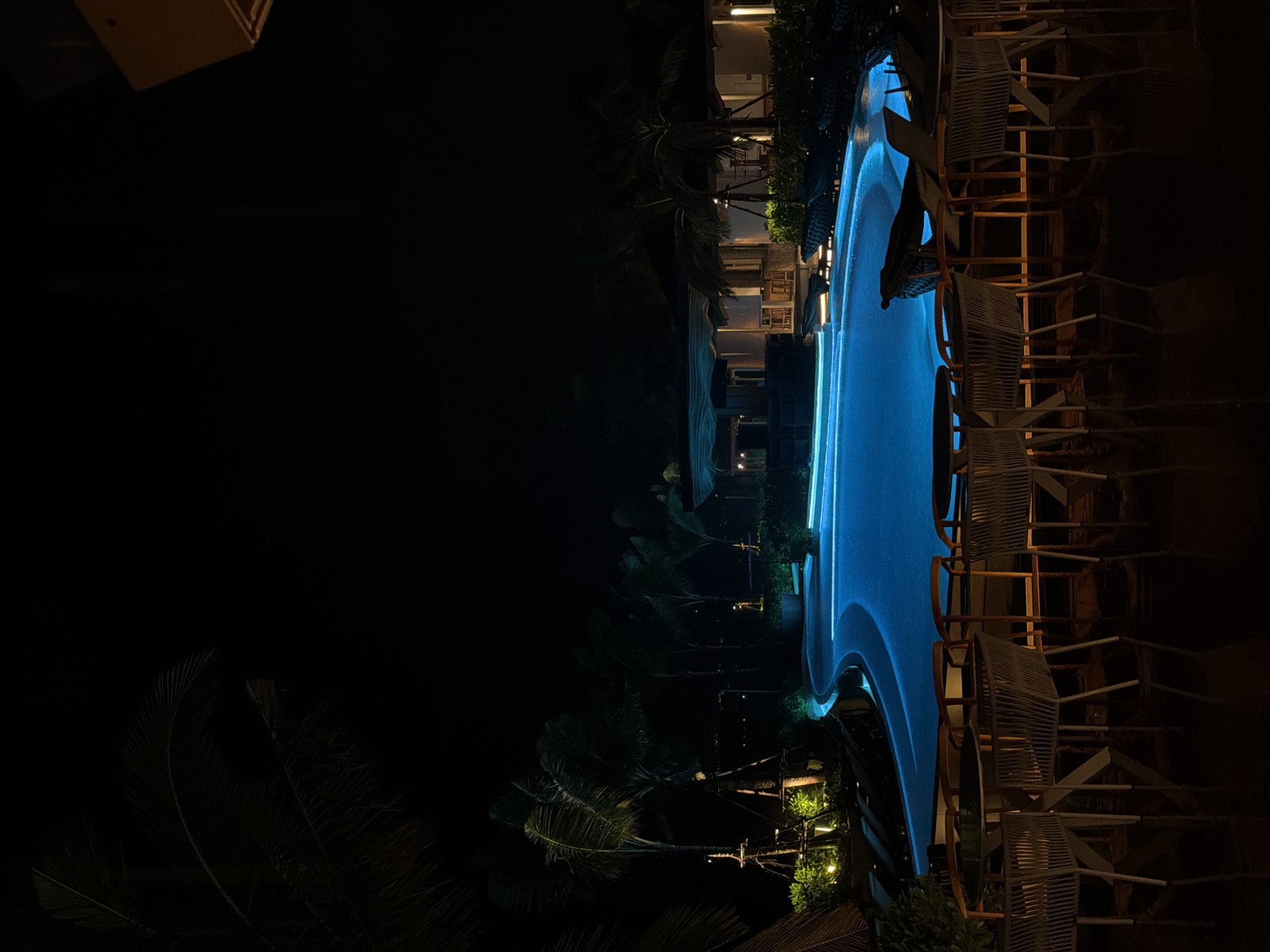 The image captures an illuminated swimming pool at night in Choeng Thale, Thailand. The pool is a bright blue color and is surrounded by several chairs and tables. Palm trees and other foliage are visible around the pool area. Buildings with lights are also present in the background. The scene is dark, with the pool and surrounding lights providing the main source of illumination.