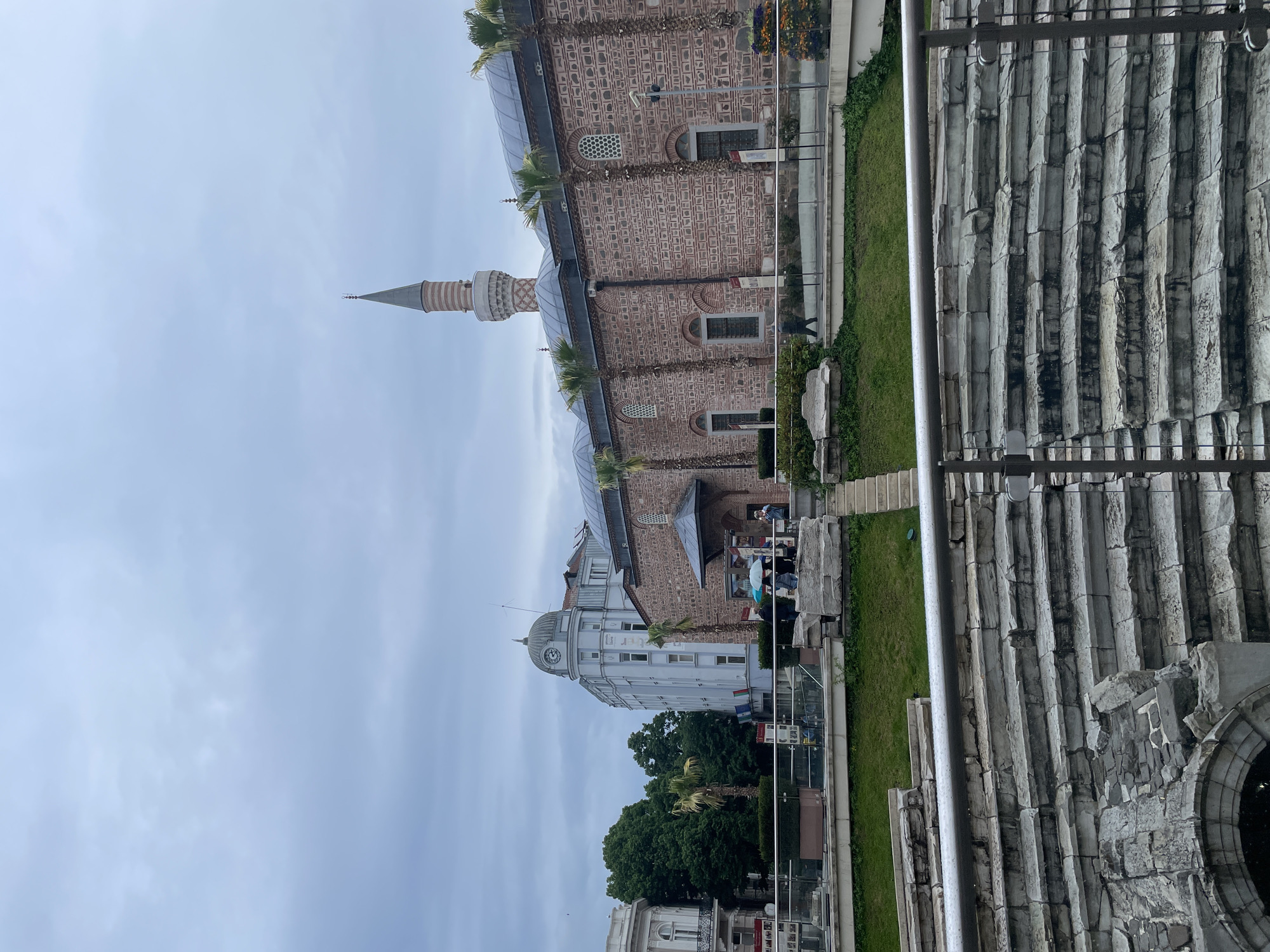 The image shows a view of the city of Plovdiv, Bulgaria. Visible in the shot are the Dzhumaya Mosque, an Ottoman-era mosque, and the Roman Stadium ruins. The mosque is brick-built with a tall minaret. The Roman Stadium, in the foreground, is characterized by its gray stone pillars and arches. Some people can be seen near the mosque, possibly tourists. The sky is overcast.