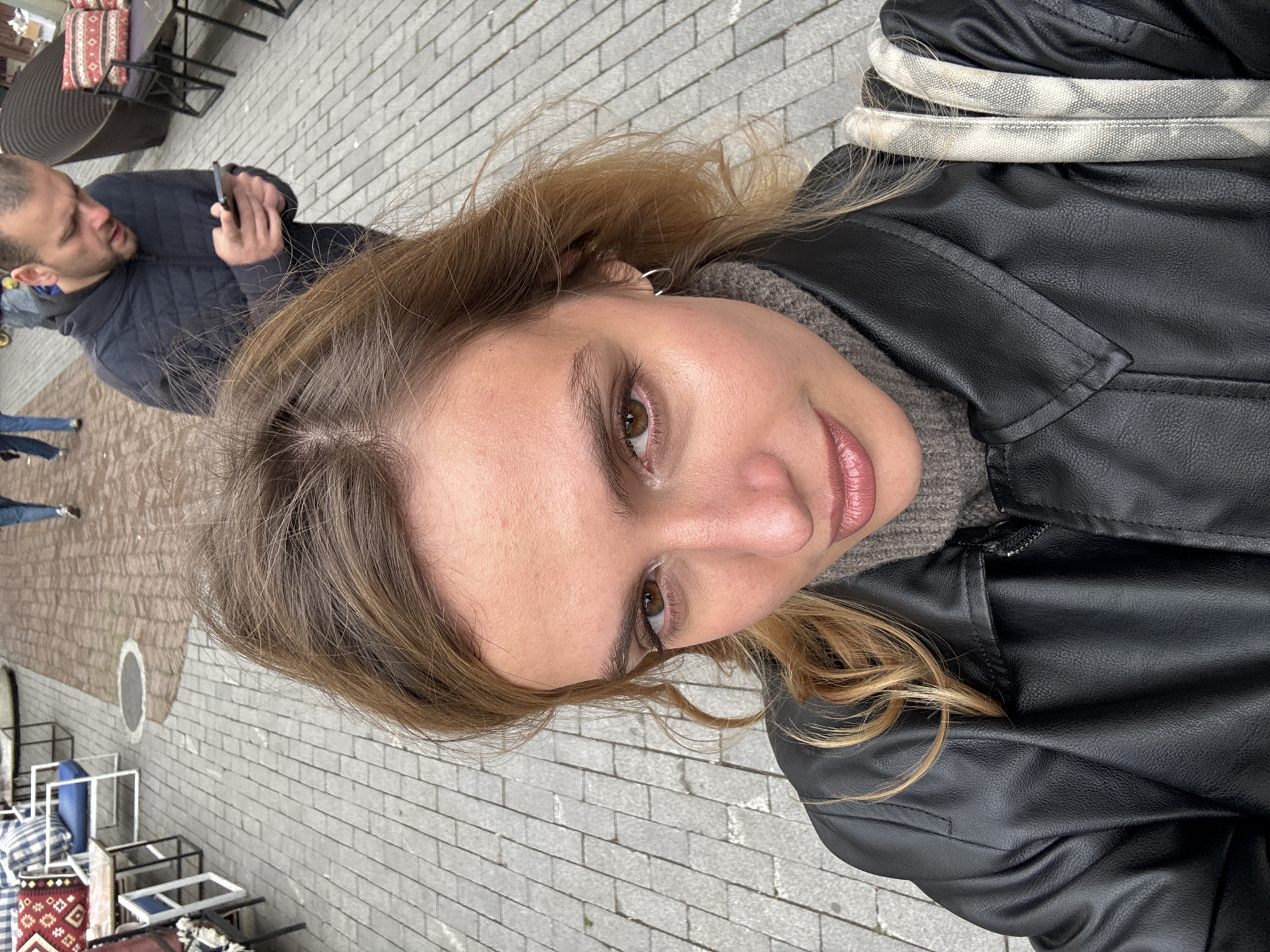 A selfie taken outdoors, showing a woman from a high angle, wearing a black leather jacket over a gray sweater. She has light brown hair with blonde highlights and light skin. Her facial features include brown eyes, fair skin, and light pink lips. In the blurred background to the left, a man is visible in a black jacket, looking at his phone. The setting appears to be a paved outdoor area with tables and chairs, potentially a café in Tbilisi, Georgia, given the style of the furniture and the paving.