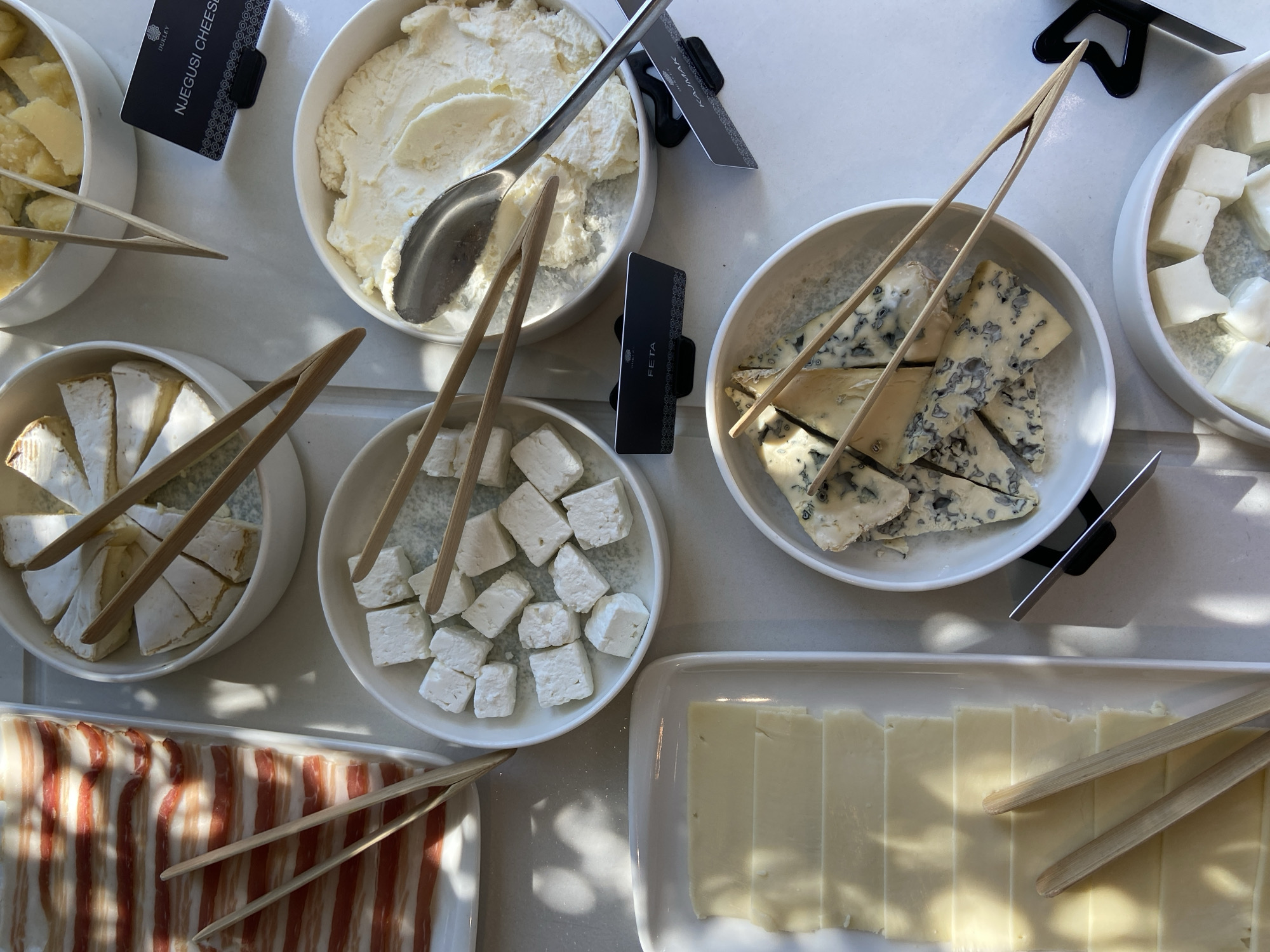 A high-angle shot depicts a cheese and meat buffet, seemingly indoors under bright natural light. The various cheeses are presented in white bowls and trays. From left to right: a bowl of sliced yellow cheese with a black label that reads "NJEGUSI CHEESE"; a bowl of white, creamy cheese with a spoon; a bowl of sliced brie; a bowl of cubed white cheese with a label that says "FETA"; a bowl of sliced blue cheese; a bowl of cubed white cheese; a rectangular dish of sliced cheese; and a tray of streaky bacon. Each cheese and meat offering has a set of wooden tongs. The scene appears to be a high-end buffet, potentially in a hotel or restaurant in Budva, Montenegro, given the types of cheeses and meats commonly found in that region. No people are visible in the image.