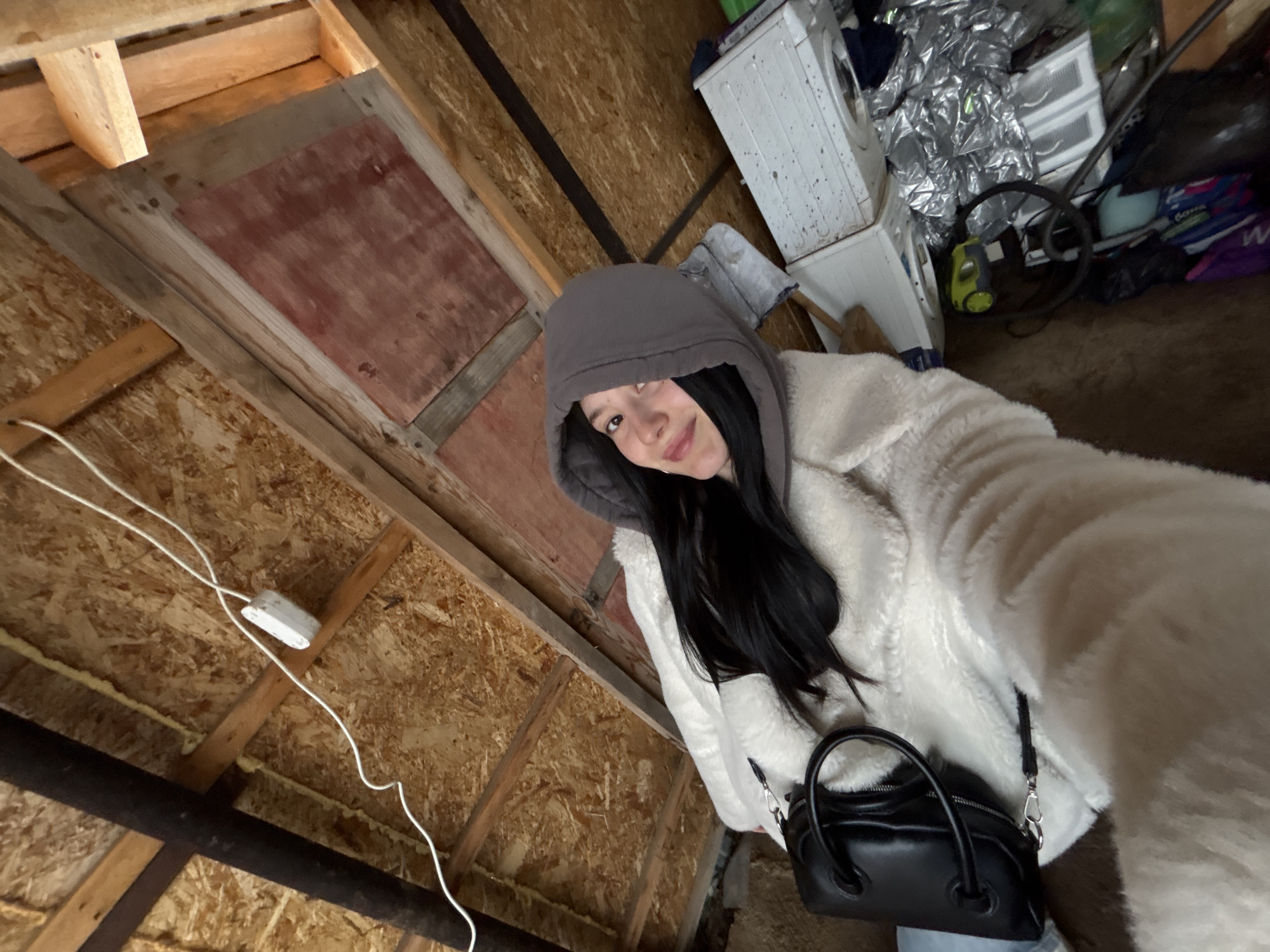 A person in a white faux fur coat and a gray hoodie stands in a cluttered attic-like space. The walls and ceiling are made of wood, possibly oriented strand board (OSB). A white electrical box and wiring are visible on the ceiling. Behind the person are various items including a washing machine, foil-wrapped items, and a vacuum cleaner. The floor appears to be stained. The person is holding a small black handbag. The photo appears to have been taken at an angle. It is impossible to ascertain whether this image was taken in the city of Фоки, Russia from visual information.