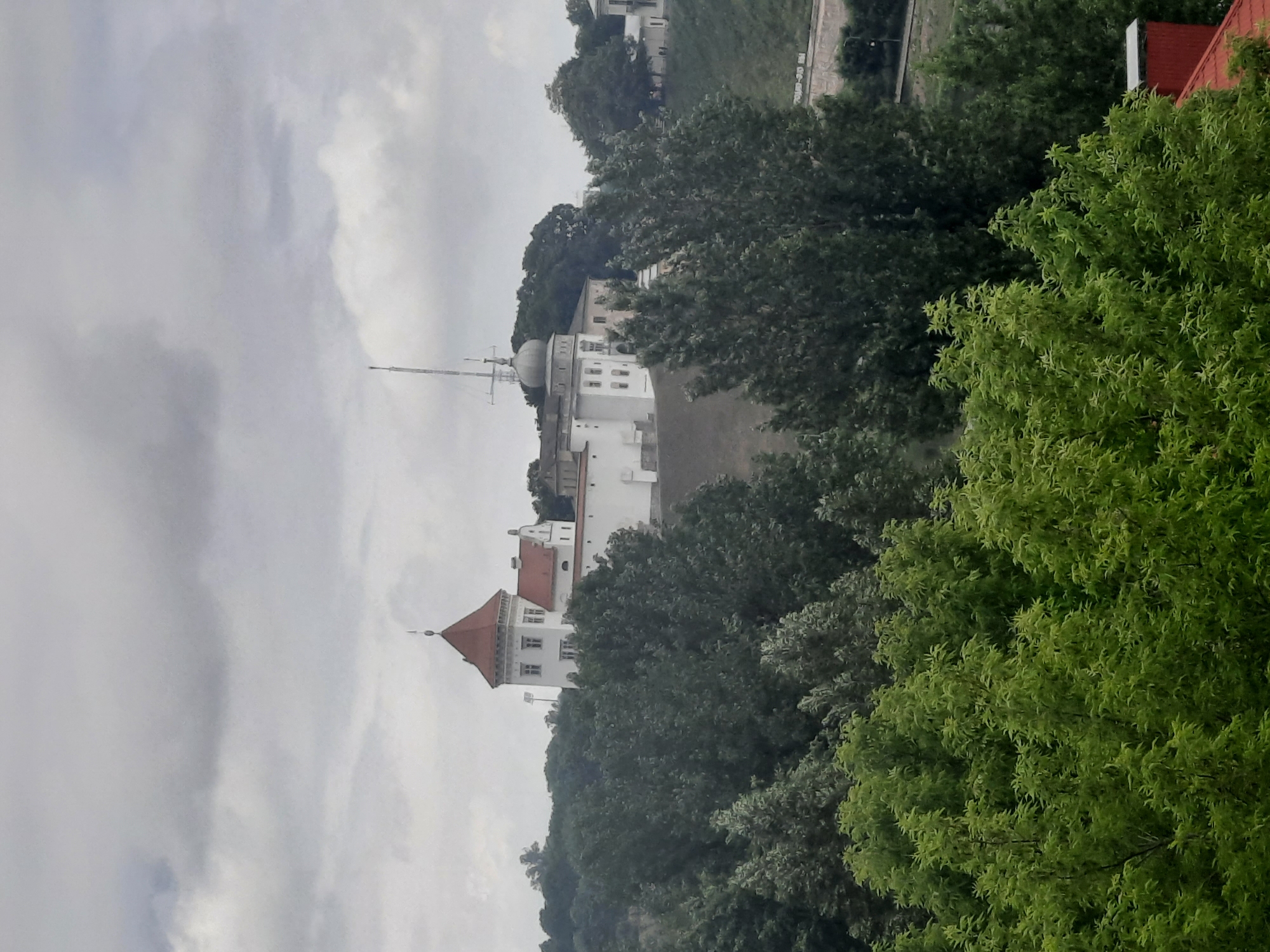 The image shows a white building with a red-tiled roof, possibly a castle or palace, situated on a hill. The building has multiple windows and a distinctive domed tower topped with a spire. It is surrounded by lush green trees. The sky is overcast with gray clouds. In the foreground, there are more trees, with some showing bright green foliage. The scene appears to be a daytime view from a slightly elevated perspective, possibly overlooking a park or a historical site within the city of Hrodna, Belarus. The lower right corner of the image shows a glimpse of a red-tiled roof, suggesting the presence of other buildings nearby. There are no people visible in the image.