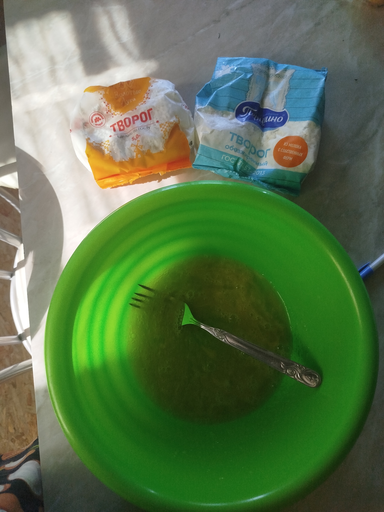 The image depicts a bright green mixing bowl containing a yellow, liquid mixture, with a silver fork resting inside. Two packages of cheese curd, labeled in Russian as "Творог," are positioned behind the bowl. The package on the left is predominantly orange and white, while the one on the right is blue and white. The scene is set on a light-colored surface, possibly a table, with shadows cast from an unseen light source. The overall composition suggests food preparation. The image does not provide any specific information about the city of Zavodoukovsk, Russia, or any people.
