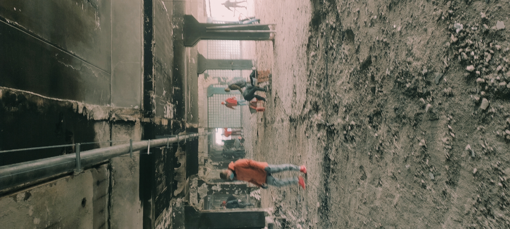 This image depicts a large, industrial-style interior space, possibly an abandoned or repurposed facility, located in Fryazino, Russia. The perspective is from a slightly elevated vantage point, looking down into a long, cavernous area.
On the left, a massive, dark, and heavily weathered concrete or metal wall dominates, showing signs of significant age, staining, and wear. A large, grey metallic pipe runs horizontally along the lower section of this wall, supported by brackets, with a thin railing above it.
The right side of the image reveals a rough, uneven ground surface composed of dirt, gravel, and scattered debris or broken concrete, suggesting a raw or undeveloped floor.
Several figures are visible within the space, indicating human activity. In the foreground, a person in an orange jacket and blue jeans, with bright red shoes, walks across the rough ground. Further into the scene, other blurred figures are present, including one in a red jacket and another in darker clothing, suggesting movement and depth within the expansive area. Their activity appears to be walking or exploring.
In the background, more industrial structures can be discerned, possibly large concrete pillars and sections that resemble windows or skylights composed of glass blocks, allowing a muted light into the space. The overall atmosphere is stark and industrial, with a muted color palette dominated by greys, dark browns, and desaturated blues and oranges. There is no discernible text visible in the image. The lighting suggests daytime, but it appears diffused, possibly due to the enclosed nature of the building or overcast conditions.