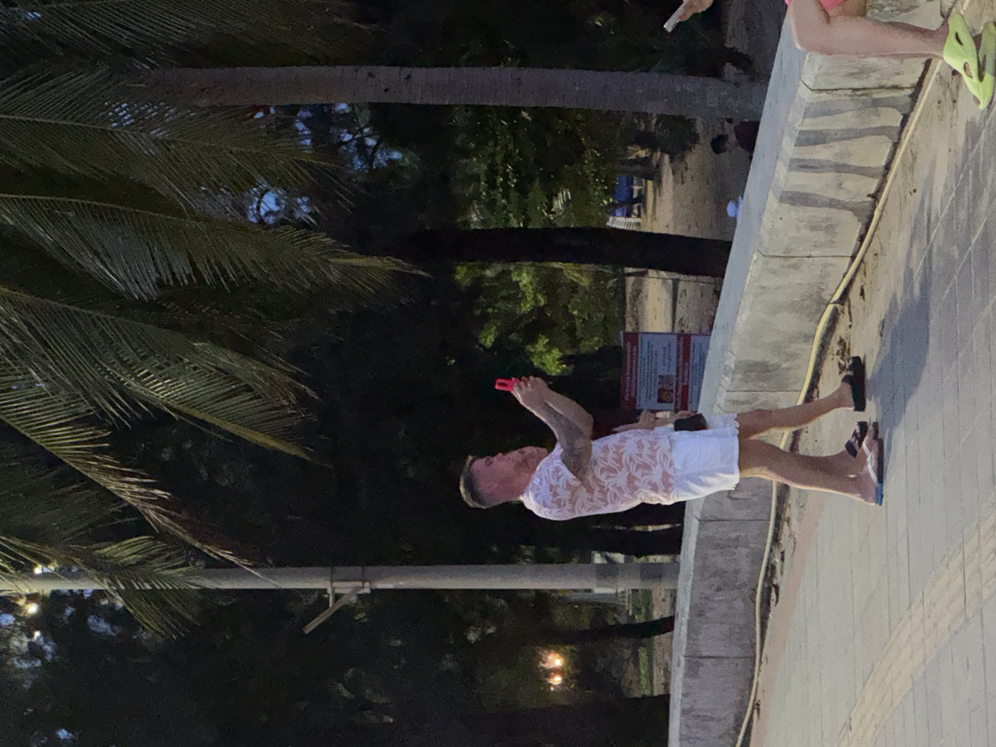 In Karon, Thailand, a man and woman stand on a paved walkway next to a concrete barrier. The man, facing left, wears a short-sleeved shirt with a palm leaf pattern and white shorts. He is holding a red smartphone in his right hand, appearing to take a photo or video. The woman, partially obscured, stands to his left, wearing white shorts. Behind them, palm trees with large fronds create a shaded canopy. A lamppost stands to the left of the man. In the background, further down the walkway and beyond the barrier, are more trees and glimpses of people and beach chairs. The lighting suggests it is late afternoon or early evening.
