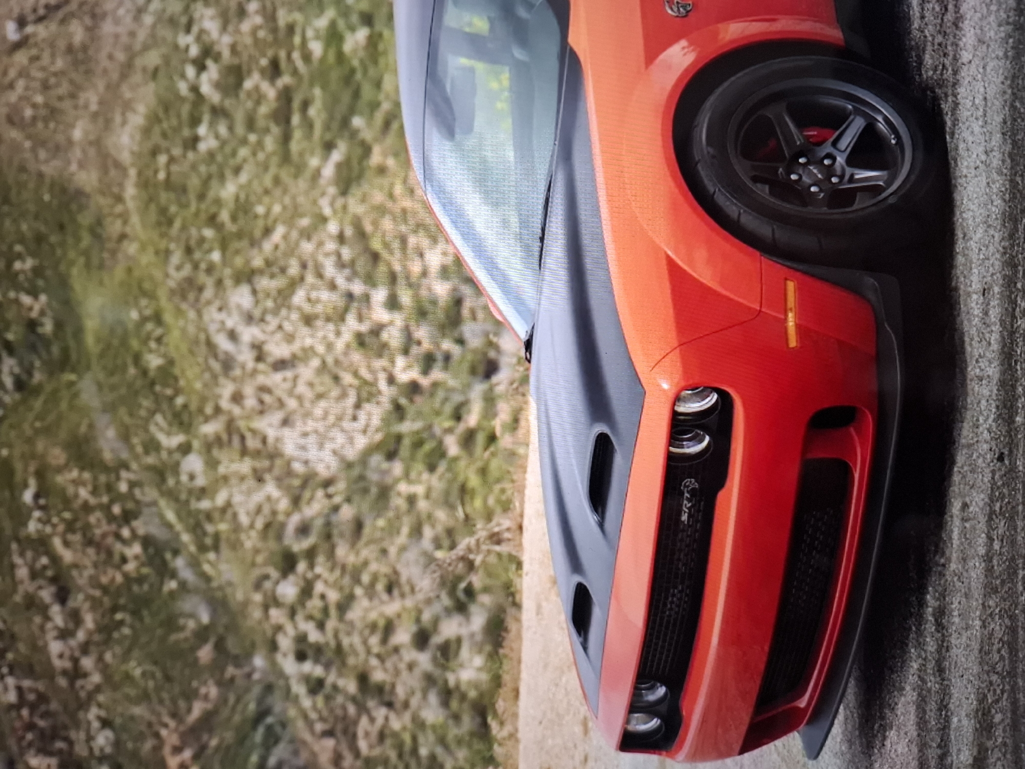 The image shows a close-up, low-angle view of the front portion of a bright orange Dodge Challenger SRT Hellcat. The car is parked on a paved surface, possibly a road or asphalt. The background consists of a dry, rocky hillside with sparse green vegetation. The car's hood is black and features prominent air intakes. The grille has "SRT" badging. The front bumper is also orange, with a black lower lip spoiler. The front wheel, visible on the right side of the frame, is black with a multi-spoke design. Red brake calipers are visible behind the wheel spokes. The windshield and part of the roof are also visible, reflecting the sky and surroundings. There are no people or other vehicles present in the image. The scene appears to be outdoors, in a somewhat arid or mountainous environment. There is no information in the image to suggest it is located in Argun, Russia.