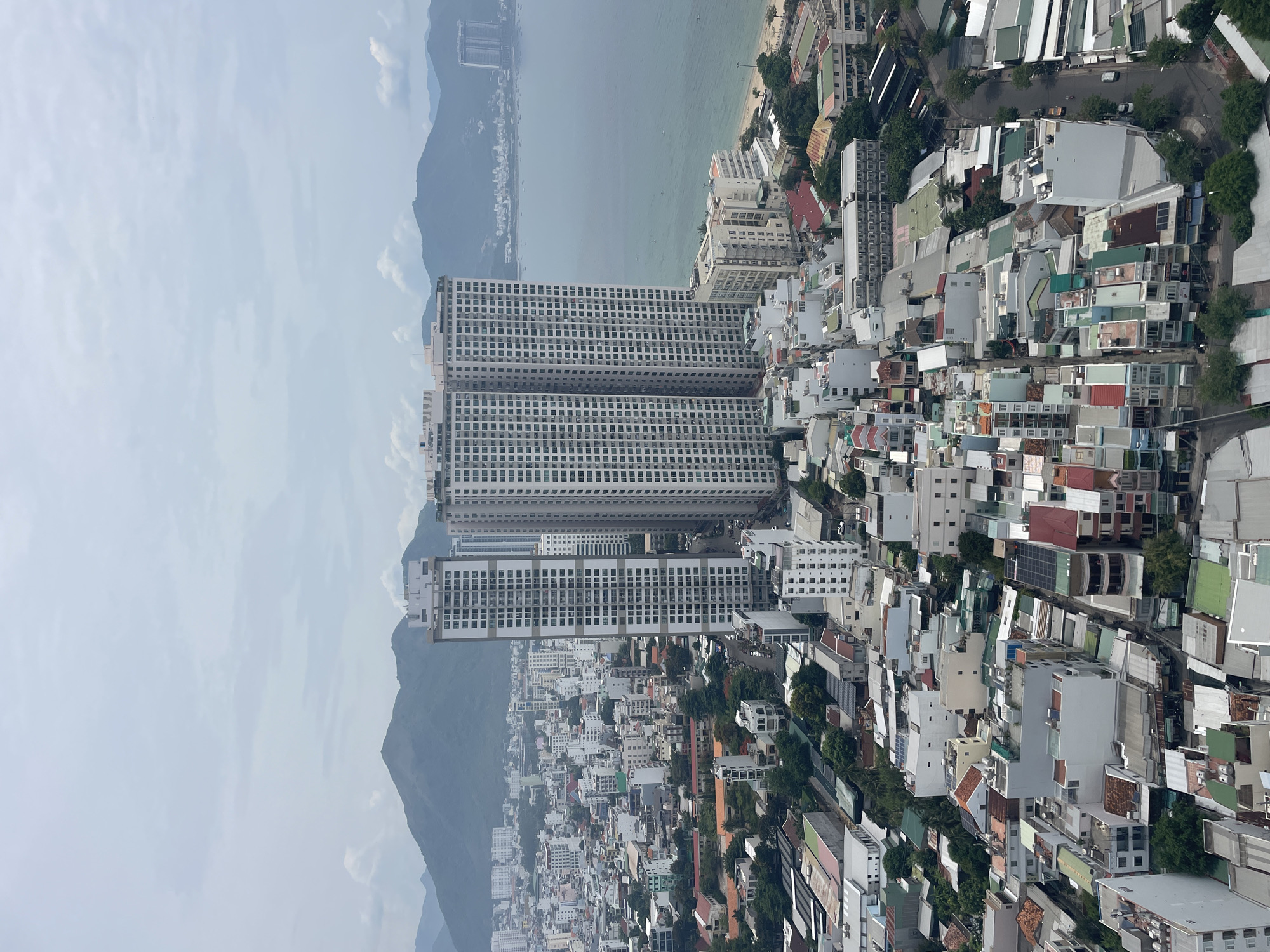 This image shows a panoramic view of Nha Trang, Vietnam, capturing the city's dense urban landscape juxtaposed with the sea and mountains. In the foreground, a cluster of multi-story residential buildings with varied roofing colors, including red, green, and grey, fills the frame. Some buildings feature visible solar panels on their roofs, suggesting renewable energy adoption. Narrow streets weave through the buildings, with a few vehicles and pedestrians visible, indicating urban activity.
Dominating the mid-ground are two towering, modern apartment complexes, characterized by their uniform white facades and numerous windows. These skyscrapers stand out against the backdrop of lower-rise buildings. Adjacent to these high-rises, a beach area is visible, with the turquoise waters of the sea gently lapping the shore.
In the background, rolling green mountains form a natural barrier, their slopes partially covered in vegetation. The sky is overcast with scattered clouds, lending a soft, diffused light to the scene. The overall impression is of a bustling coastal city with a mix of contemporary architecture and traditional housing, nestled between natural elements.