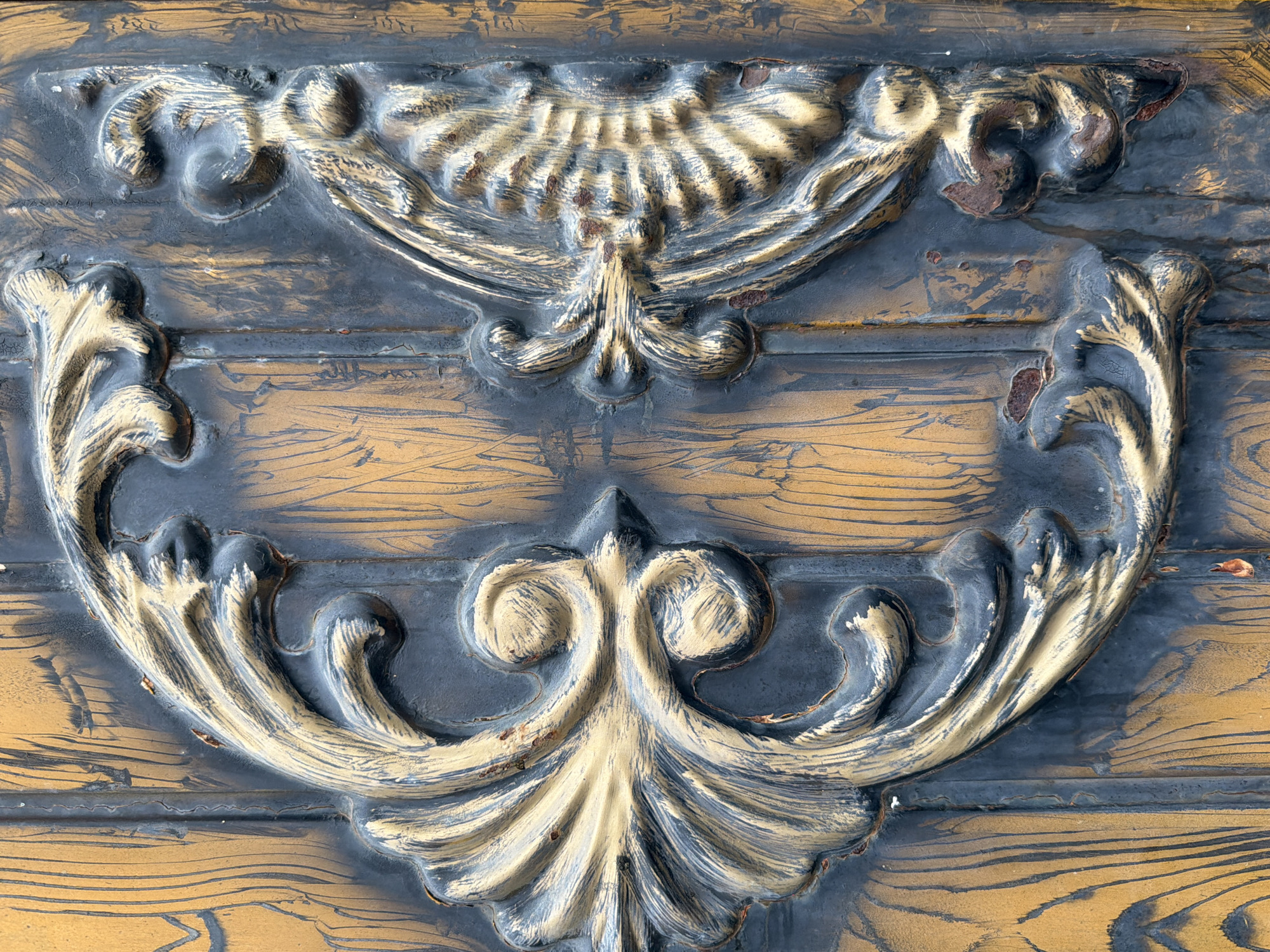 This is a close-up, high-angle, eye-level, full-shot photograph of an ornate metal gate in Sabirabad, Azerbaijan. The gate has a dark, textured surface that appears weathered and possibly rusted in places, with prominent golden-bronze decorative elements. The golden accents are stylized and resemble large leaves or floral motifs, with some appearing to have a fan-like or feathery texture. The overall impression is one of aged, decorative metalwork, likely on a building entrance or fence. The background is a textured, wood-grain-like pattern in shades of brown and gold, which could be the surface of the gate itself or a wall behind it. The lighting is natural and appears to be daylight, casting soft shadows that highlight the relief of the metalwork. There are no people or animals visible in the image.
