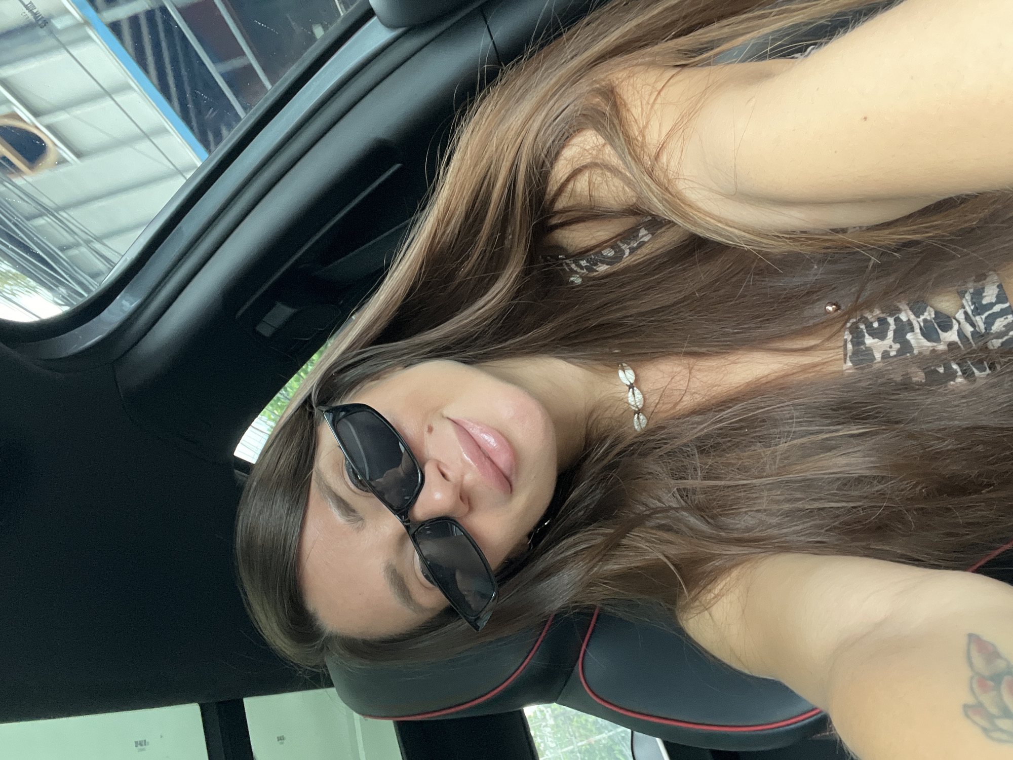 A woman is featured in a close-up, tilted selfie taken inside a car. She has long, flowing brown hair, which covers parts of her shoulders and the car seat. Her face is turned slightly towards the camera, wearing dark, rectangular sunglasses positioned a little askew, allowing a portion of her right eye to be seen. Her lips are glossy and slightly parted, suggesting a relaxed expression. Around her neck, she wears a necklace made of small white shells. She is dressed in a top with a leopard print pattern, visible beneath her hair.
The setting is the interior of a car, evidenced by the black car seat with red trim, and the car's ceiling structure. A bare arm, possibly her right, is visible on the lower right side of the frame, revealing a tattoo that appears to be a floral or lotus design. Through the car's window or sunroof, reflections of modern buildings with glass facades and the bright sky are visible, indicating it is daytime. The bright, natural lighting suggests sunny weather. The reflections hint at an urban or developed area, fitting generally with the Baan Hua Thanon, Thailand location context. Several faint, illegible white symbols or text are barely visible on a distant, reflected surface. The overall mood is casual and relaxed.