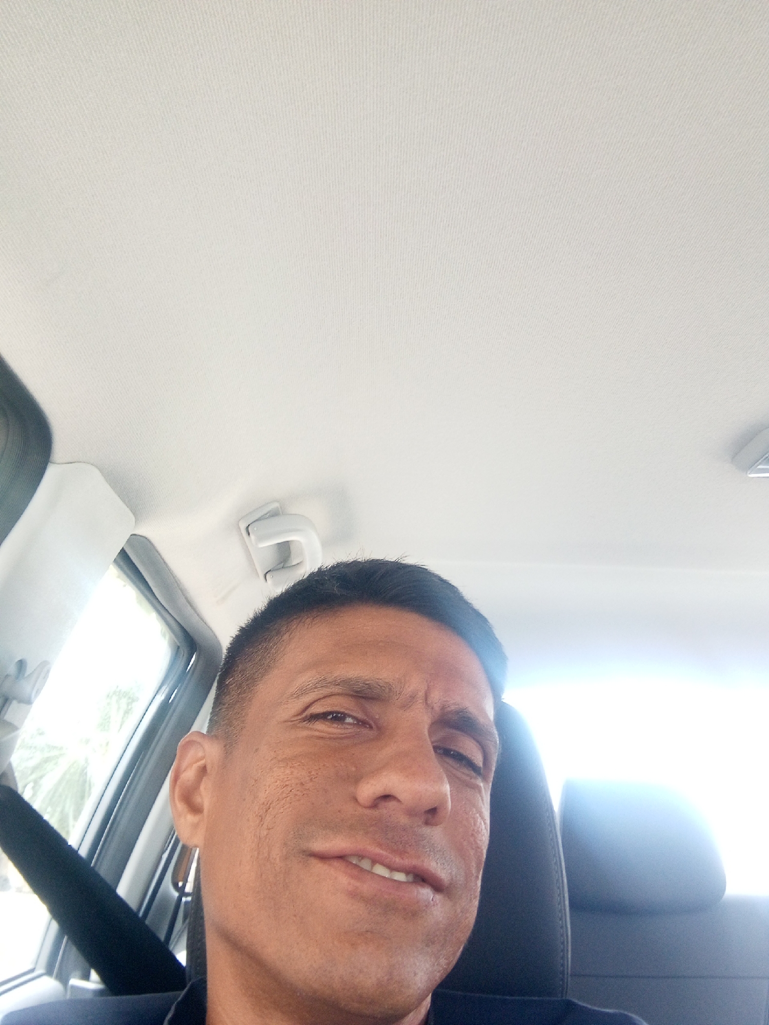 A man sits in the driver's seat of a car. He is wearing a seatbelt. The car's interior is light-colored, with a white ceiling. The man has dark hair and a light complexion. Outside the car window, it is bright and there is partial foliage. The exact location within Porlamar, Venezuela, cannot be determined from the provided image.