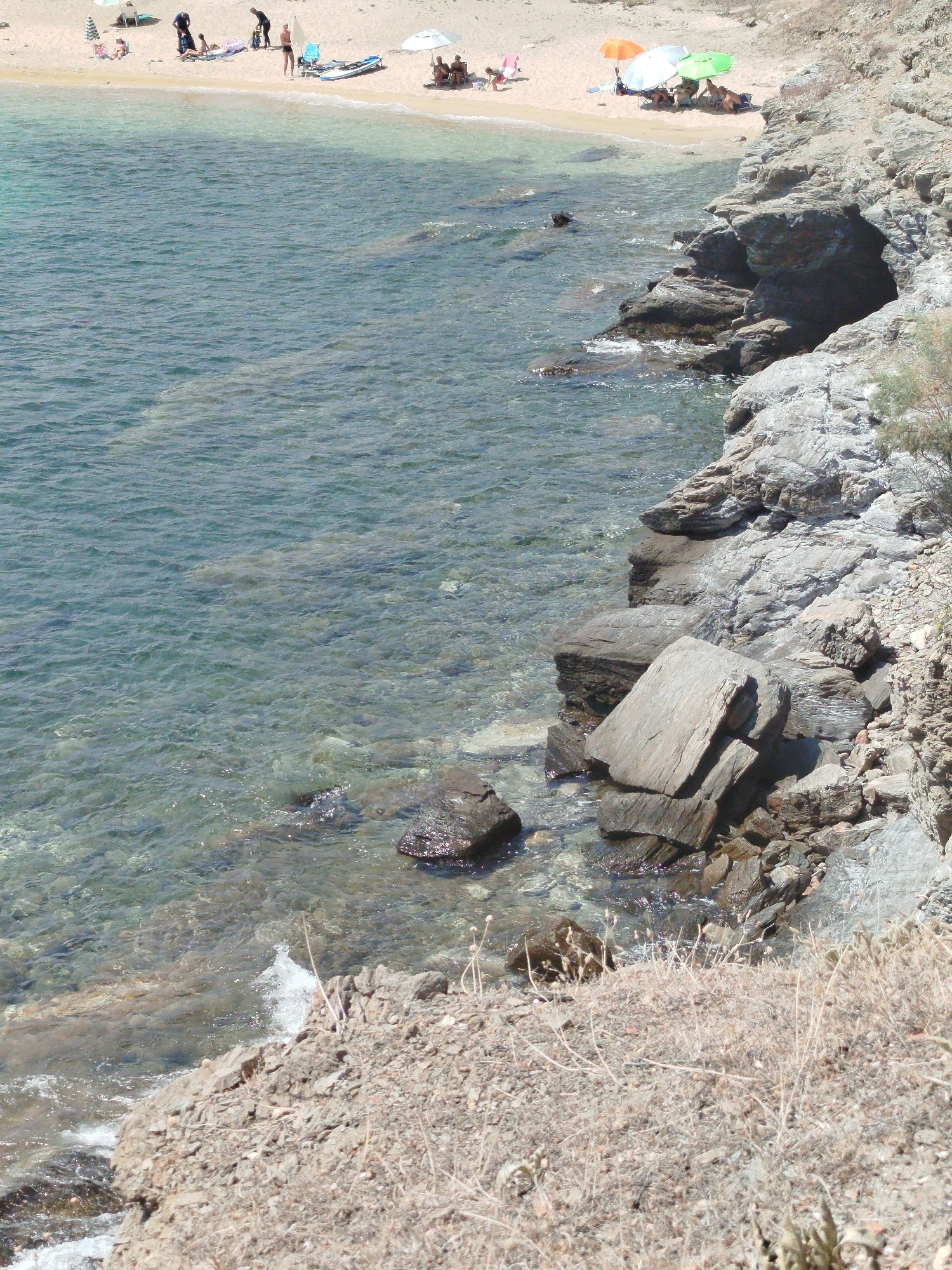 The image shows a rocky coastline with clear turquoise water. The water is shallow near the shore, revealing the sandy seabed. In the upper part of the image, a sandy beach is visible with several people sunbathing and relaxing under umbrellas. The beach is lined with vegetation. Further down the coast, there are large rocks and cliffs. The overall scene depicts a tranquil summer day at a coastal resort. The specific location is not identifiable within the image.