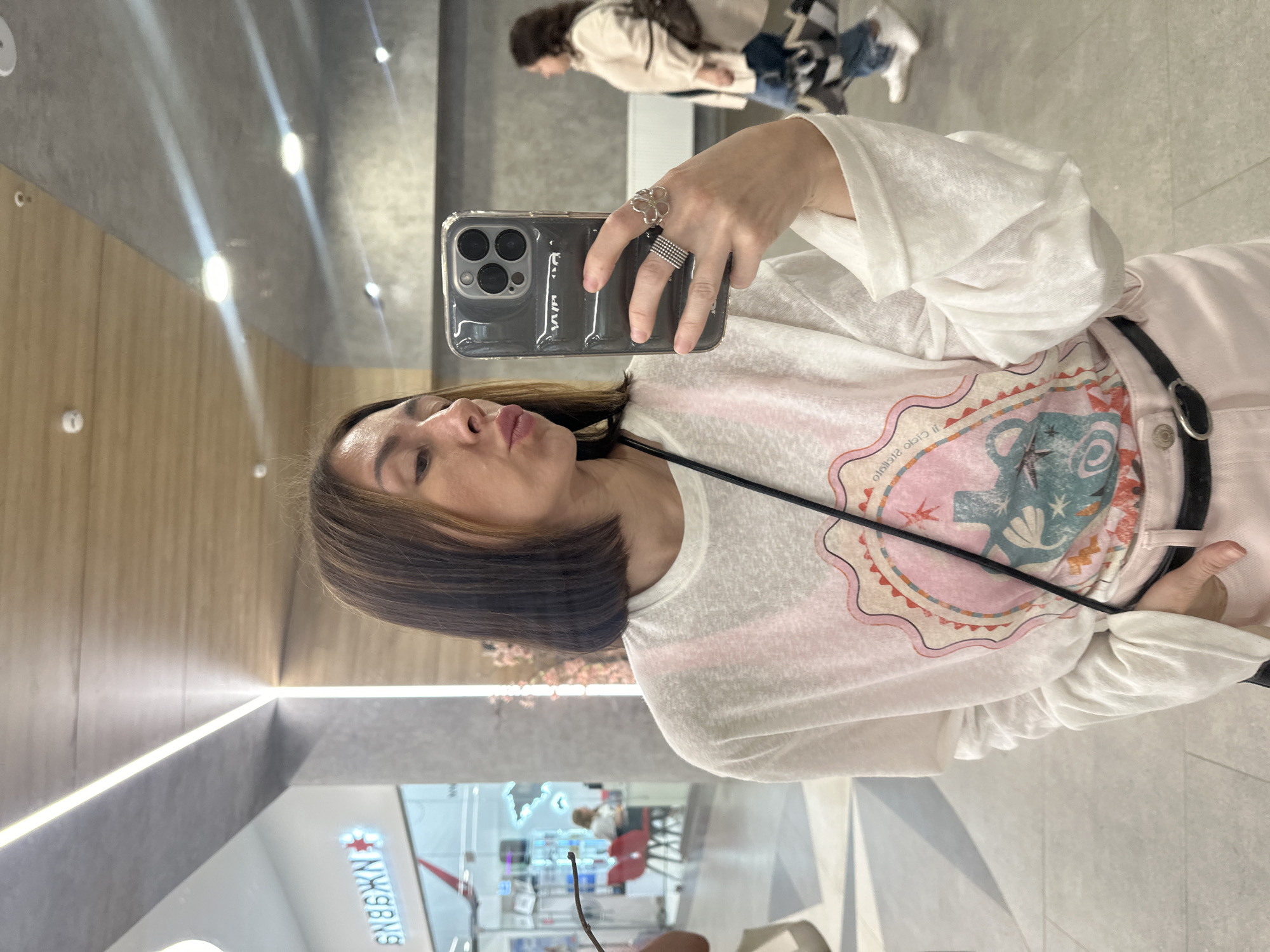 The image is a selfie taken in a brightly lit, modern shopping mall in Izhevsk, Russia. The subject is a woman with shoulder-length dark hair, wearing a white long-sleeved t-shirt with a graphic print and pink pants. She is holding a smartphone in her right hand, taking the picture, and is wearing a ring on her index finger. In the background, a blurred figure of another woman in a light-colored jacket and jeans is walking away. The mall features illuminated ceiling lights and a wooden-paneled ceiling, with a shop sign reading "SNBGEN" in Cyrillic visible on the left. The flooring appears to be light-colored tiles.